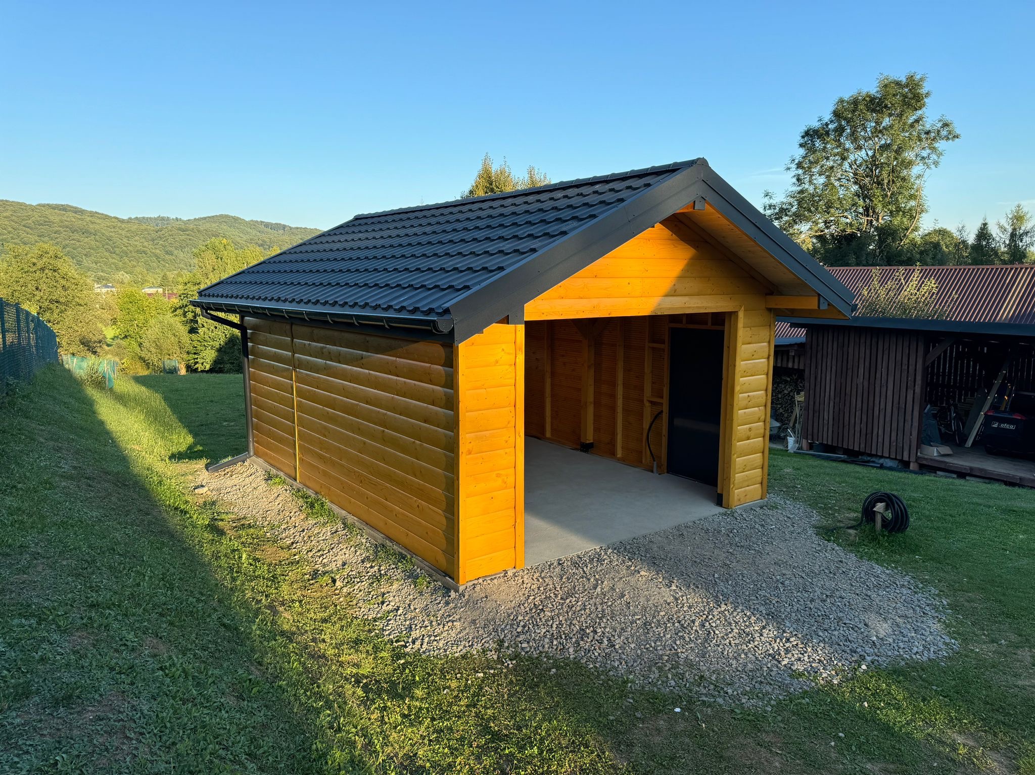 Żółta, drewniana konstrukcja garażowa z czarnym dachem, usytuowana na żwirowym podłożu, w tle zielone wzgórza i drzewa.