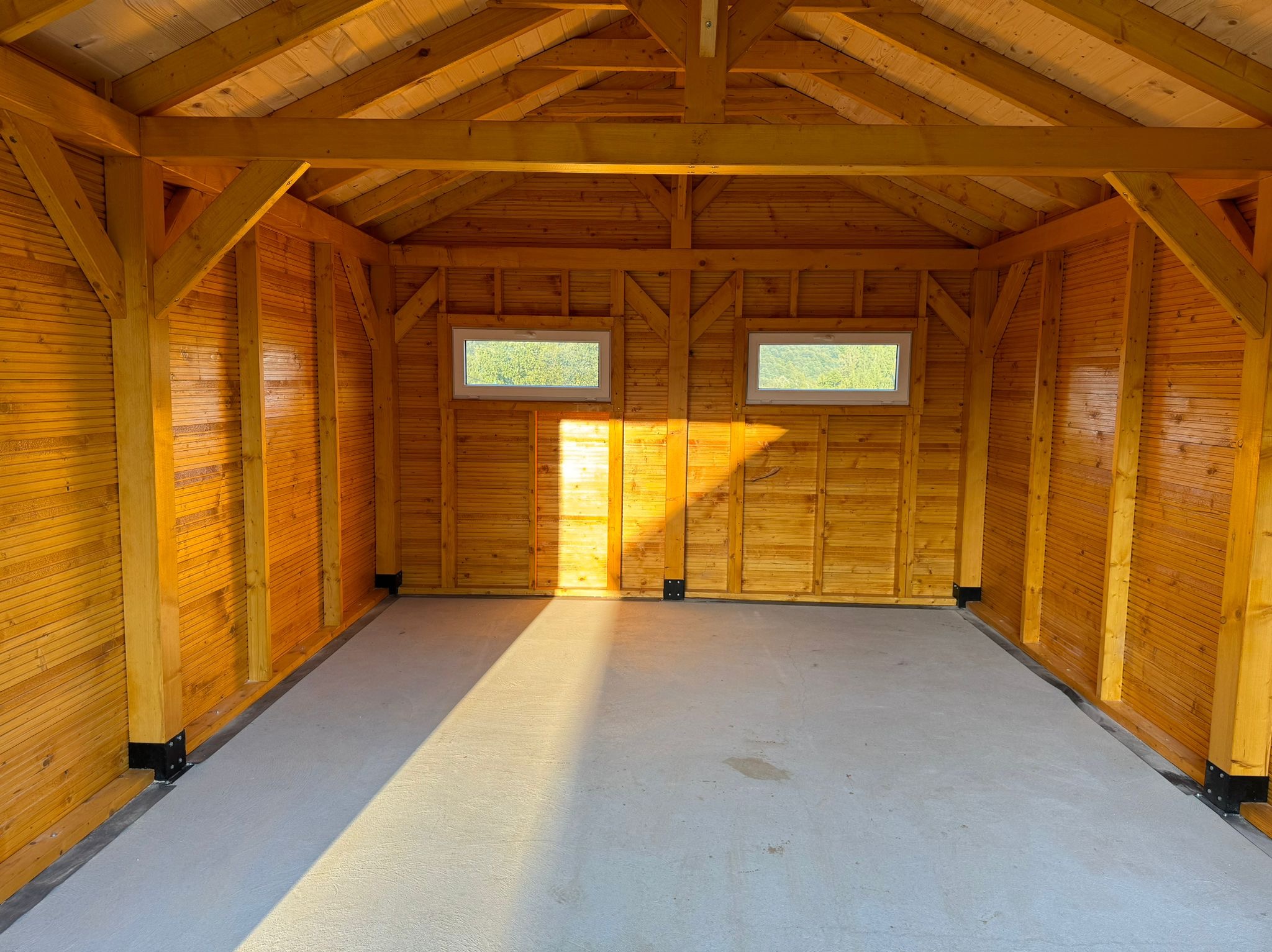 Wnętrze nowej, drewnianej konstrukcji z dwoma oknami, betonową posadzką i widocznymi elementami więźby dachowej, oświetlone wpadającym światłem słonecznym.