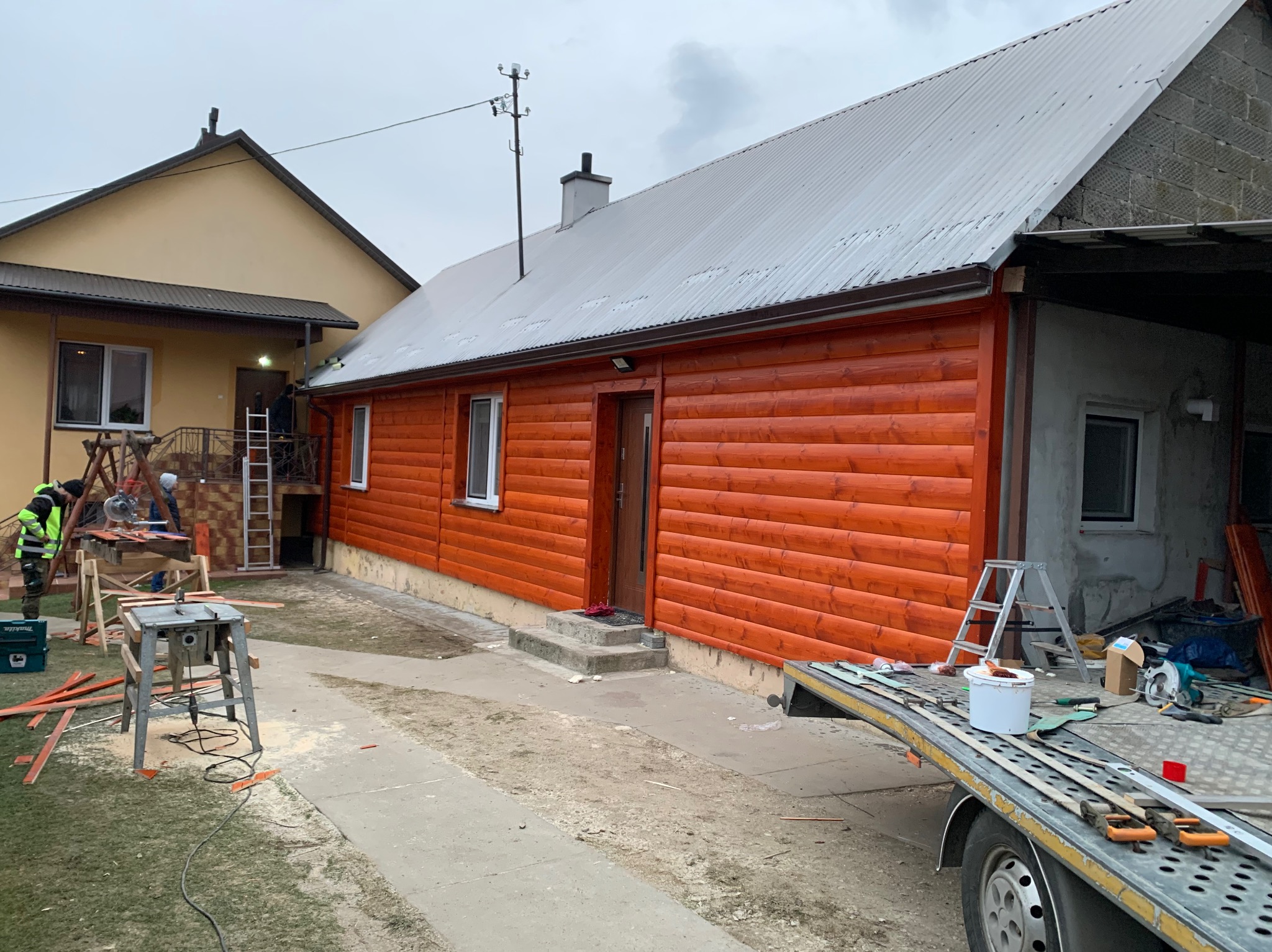 Dom z nową, pomarańczową elewacją drewnianą, widok z zewnątrz podczas prac wykończeniowych; w tle widoczny drugi budynek, na pierwszym planie narzędzia i przyczepa.