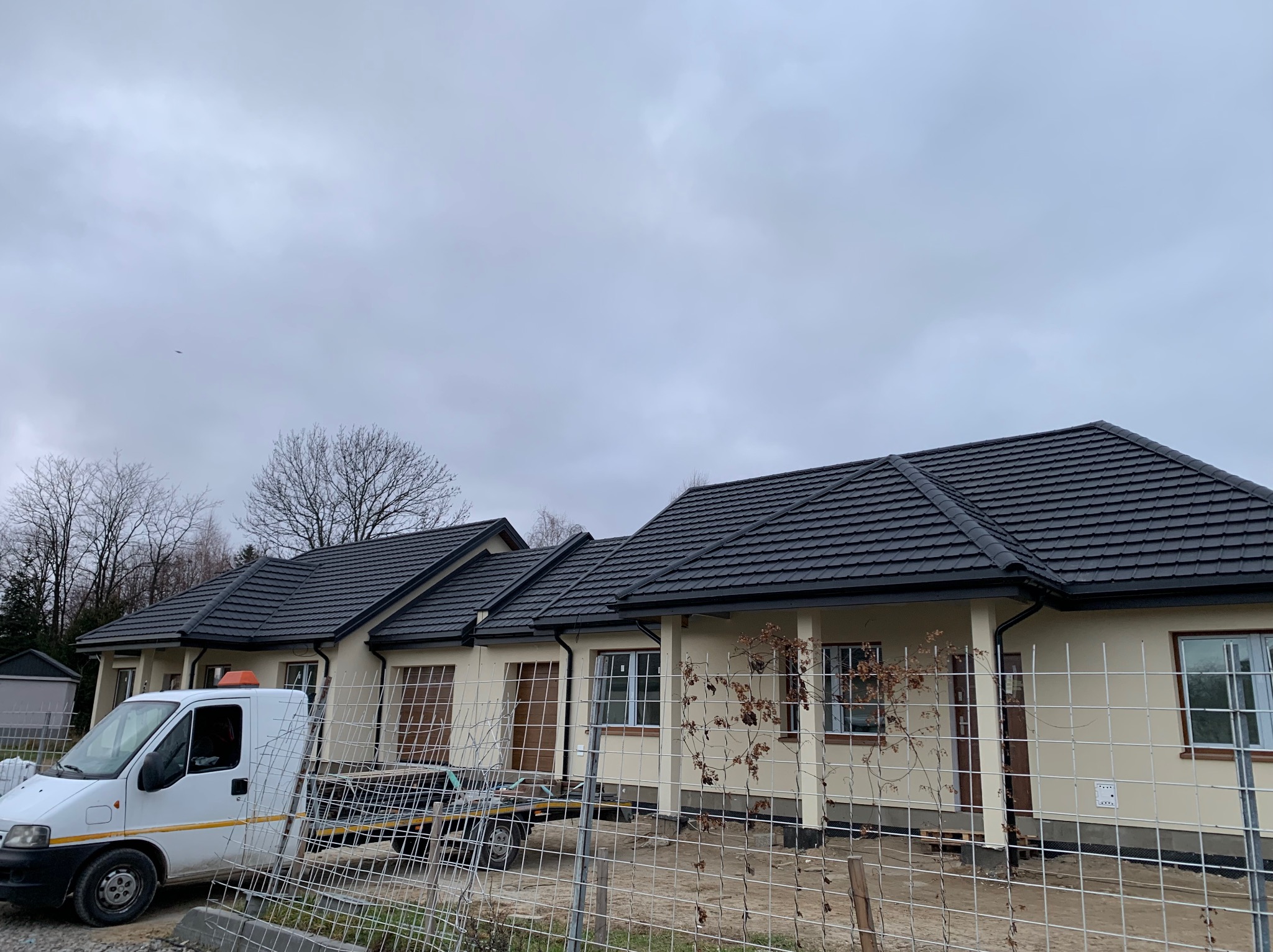 Nowo wybudowany, parterowy dom z ciemną dachówką, beżową elewacją i widocznym transportem materiałów budowlanych. Metalowe ogrodzenie tymczasowe z przodu. Pochmurne niebo w tle.