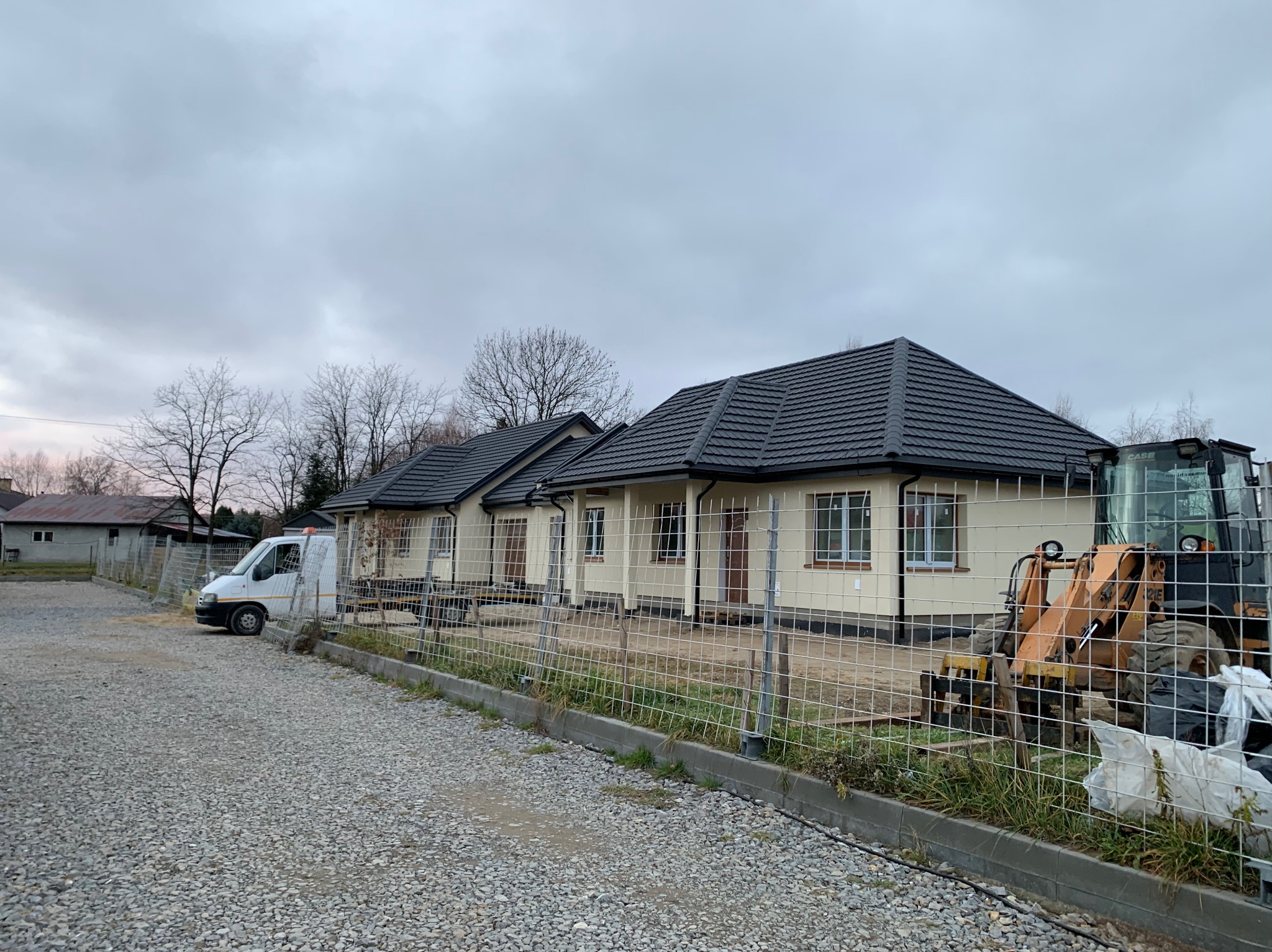 Budowa szeregu domów jednorodzinnych z ciemną dachówką, widoczny biały bus i pomarańczowa koparka za ogrodzeniem z siatki, teren wysypany żwirem, pochmurne niebo.