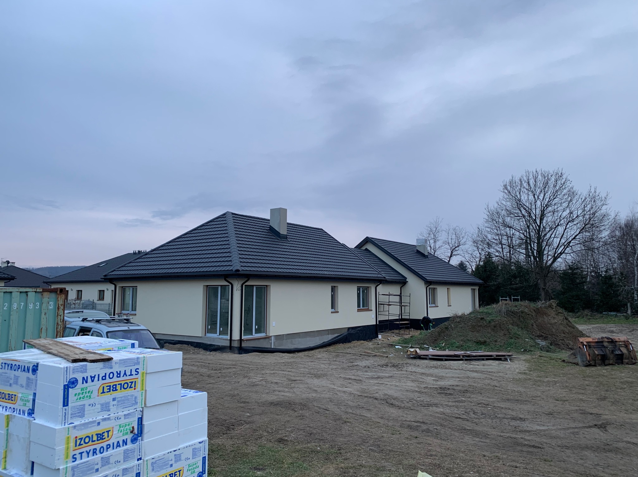 Nowy dom jednorodzinny z ciemną dachówką, częściowo ocieplony, z widocznymi paczkami styropianu IZOLBET Super Fasada 038 na pierwszym planie i stertą ziemi oraz łyżką koparki w tle.
