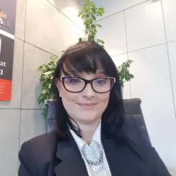 Portret uśmiechniętej kobiety w okularach i marynarce, siedzącej na krześle biurowym, z rośliną i klimatyzatorem w tle.