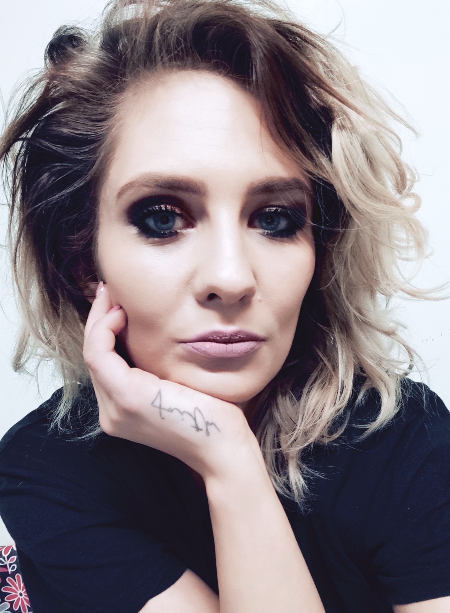 Portret kobiety z intensywnym makijażem oczu, blond pasemkami i tatuażem na nadgarstku, opierającej brodę na dłoni.