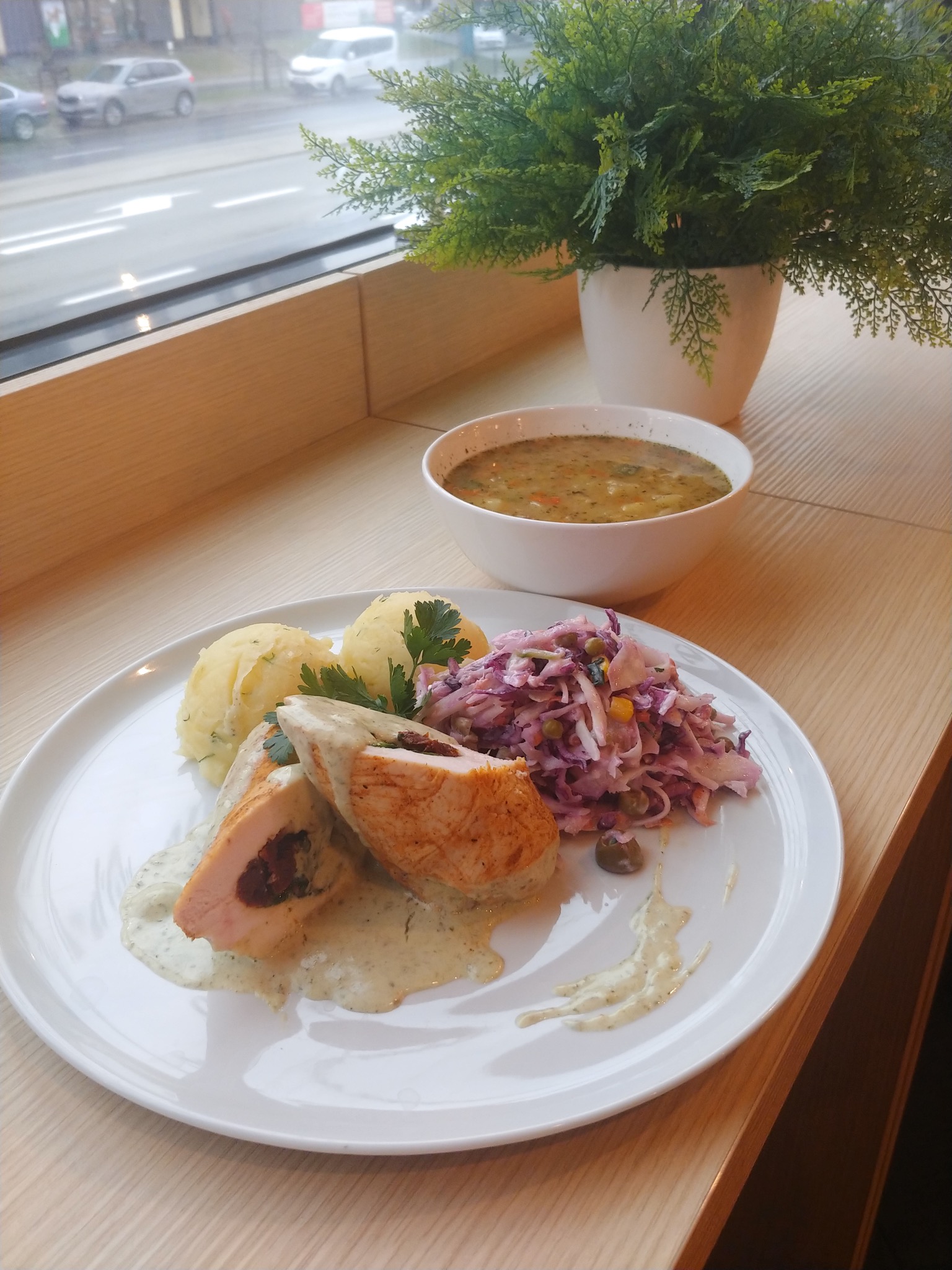 Elegancko podane danie obiadowe na białym talerzu: roladka z kurczaka z farszem, ziemniaki puree z ziołami, surówka z czerwonej kapusty i zupa w tle, ustawione przy oknie z widokiem na ulicę.