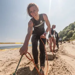 Uśmiechnięta, umorusana piaskiem kobieta w czarnym stroju sportowym ciągnie linę przymocowaną do kamienia na piaszczystej plaży podczas ekstremalnego biegu z przeszkodami, w tle inni uczestnicy...
