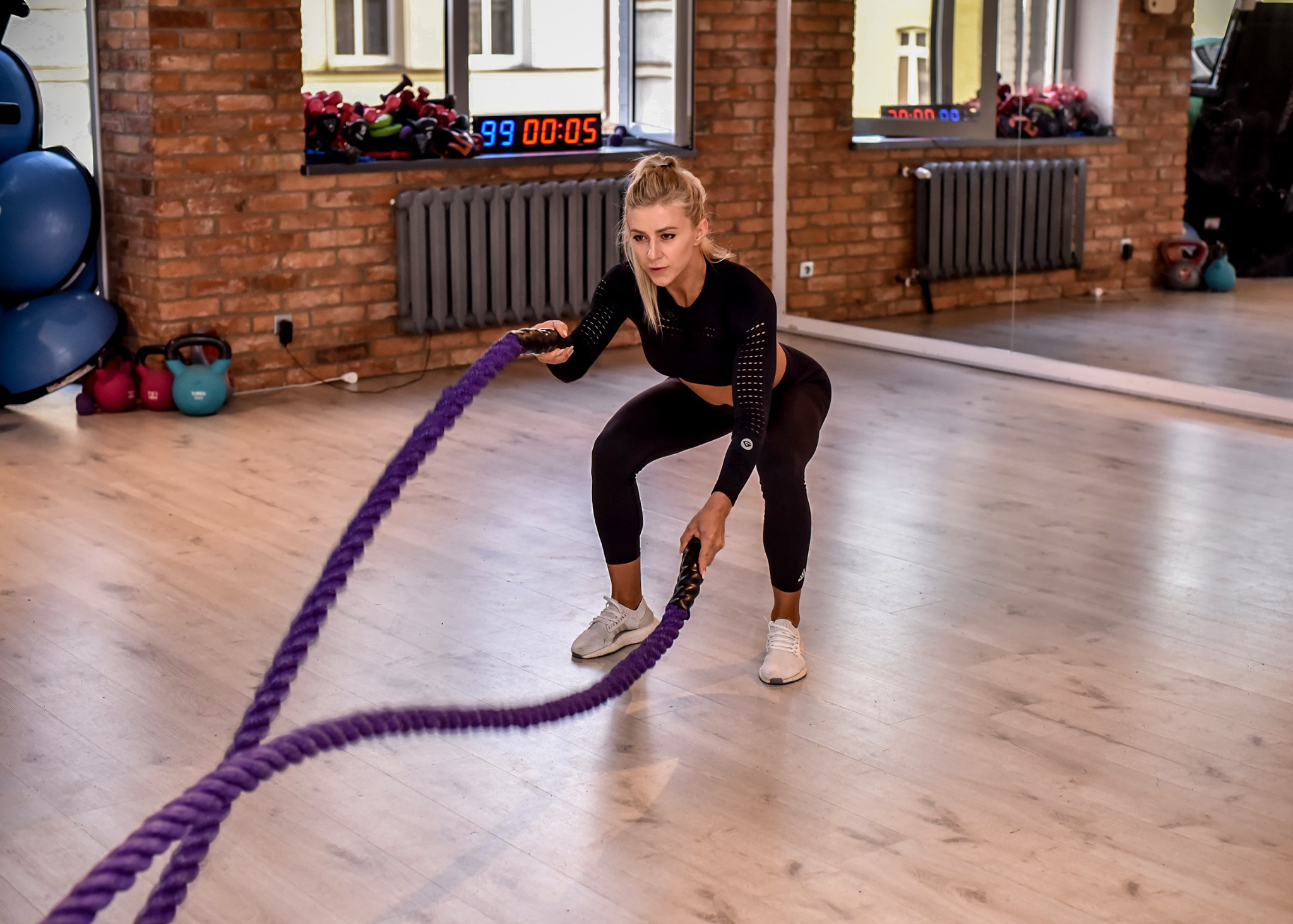 Kobieta w czarnym stroju sportowym ćwiczy z purpurowymi linami w siłowni z ceglaną ścianą i drewnianą podłogą, na ścianie widoczny elektroniczny zegar.