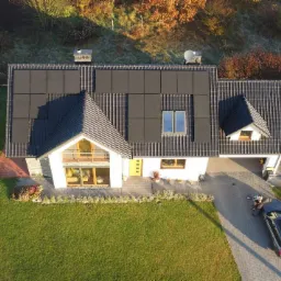 Widok z drona na nowoczesny dom jednorodzinny z panelami słonecznymi na dachu, zaparkowany samochód na podjeździe oraz zadbany trawnik i ogród.