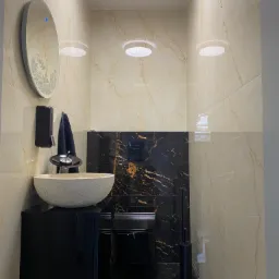 Nowoczesna toaleta z beżowymi i czarnymi marmurkowymi płytkami, okrągłym lustrem z podświetleniem LED, kamienną umywalką nablatową i czarną armaturą.