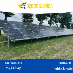 Rozległa instalacja paneli słonecznych o mocy 49,14 kWp na zielonej, wiejskiej działce, z drzewami w tle i logo firmy OZE ze Słońca w górnej części zdjęcia.
