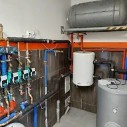 Węzeł cieplny z licznymi rurami w izolacji w kolorach niebieskim i czerwonym, zaworami, regulatorami, dwoma zbiornikami akumulacyjnymi i zasobnikiem ciepłej wody użytkowej w pomieszczeniu...