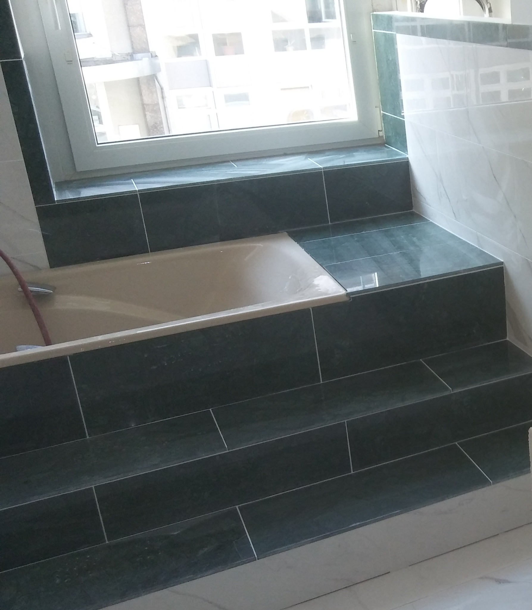 Wanna z obudową z ciemnych, błyszczących płytek ceramicznych, z widoczną baterią i fragmentem węża, obok okno i ściana z marmurkowych płytek.