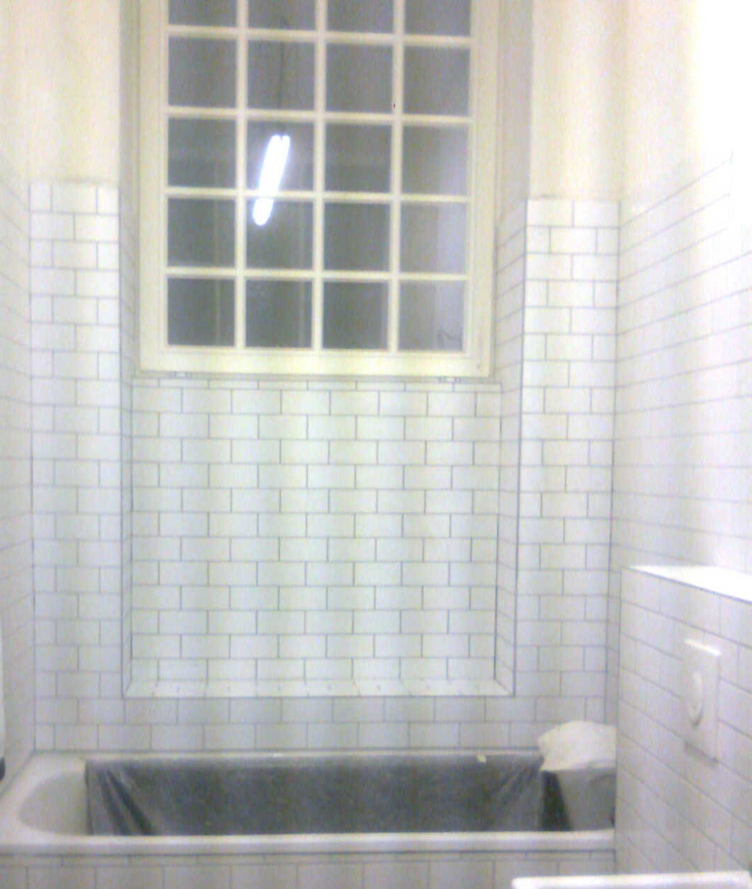 Łazienka w trakcie remontu: białe, prostokątne płytki na ścianach, okno z podziałami, wanna zabezpieczona folią.