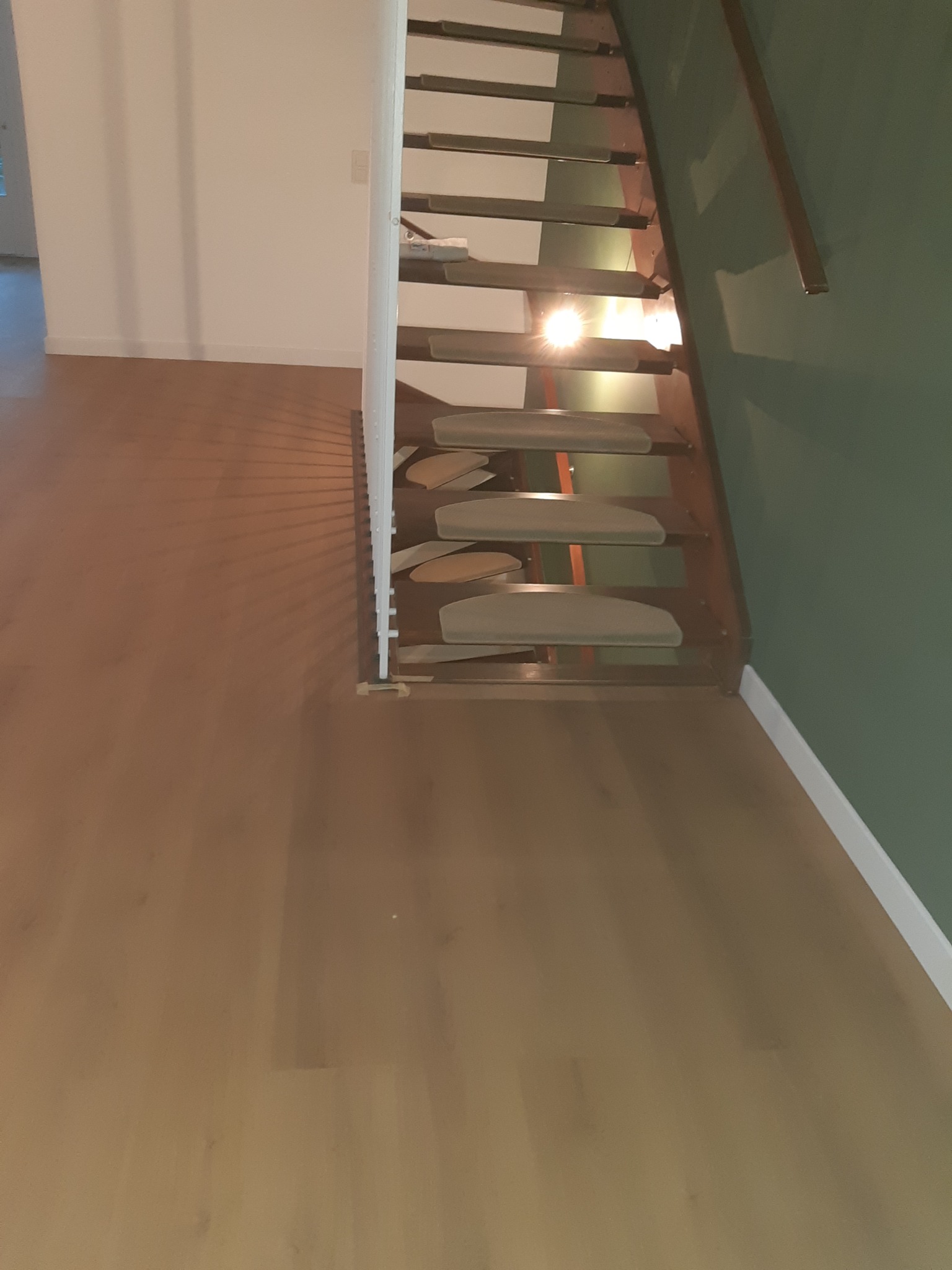 Nowoczesne, ażurowe schody z drewnianymi stopniami i metalową konstrukcją, oświetlone od spodu, na tle zielonej ściany i drewnianej podłogi.