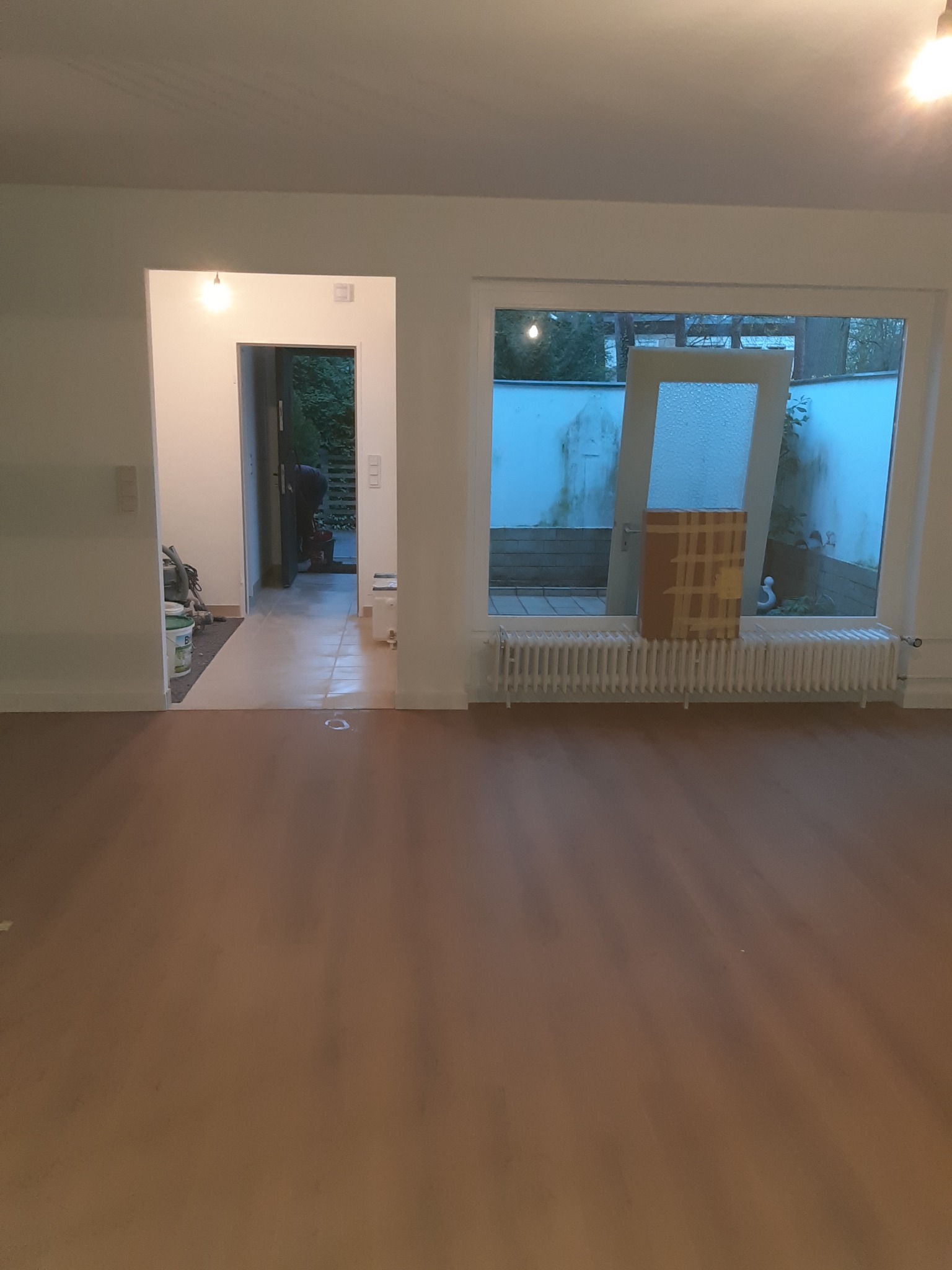 Wnętrze w trakcie prac wykończeniowych: świeżo położona, brązowa podłoga, widoczne przygotowania do dalszych prac, w głębi pomieszczenia osoba w trakcie pracy, wiadro z farbą i odkurzacz.