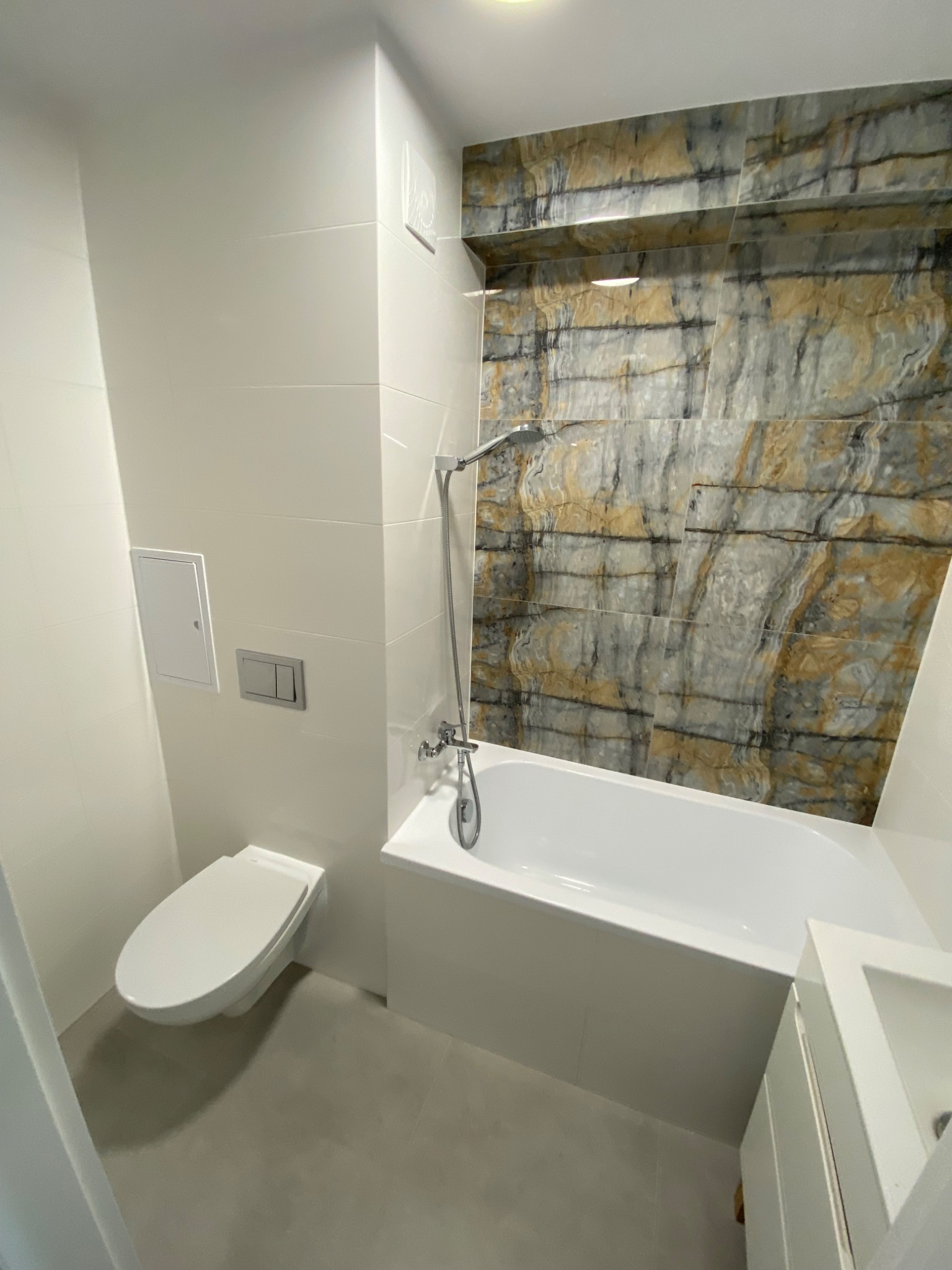 Nowoczesna łazienka z białymi płytkami, marmurową ścianą nad wanną i podwieszaną toaletą.