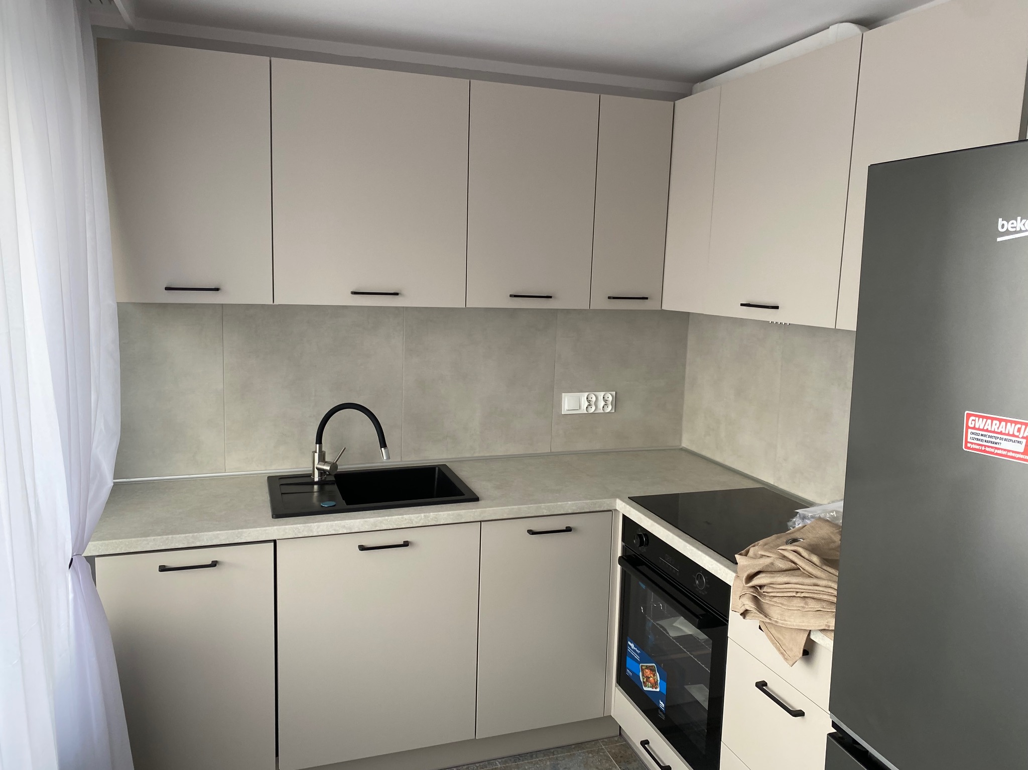 Nowoczesna kuchnia z beżowymi szafkami, czarnym zlewem i baterią, szarymi płytkami na ścianie, białą firanką w oknie i lodówką Beko z naklejką gwarancyjną.