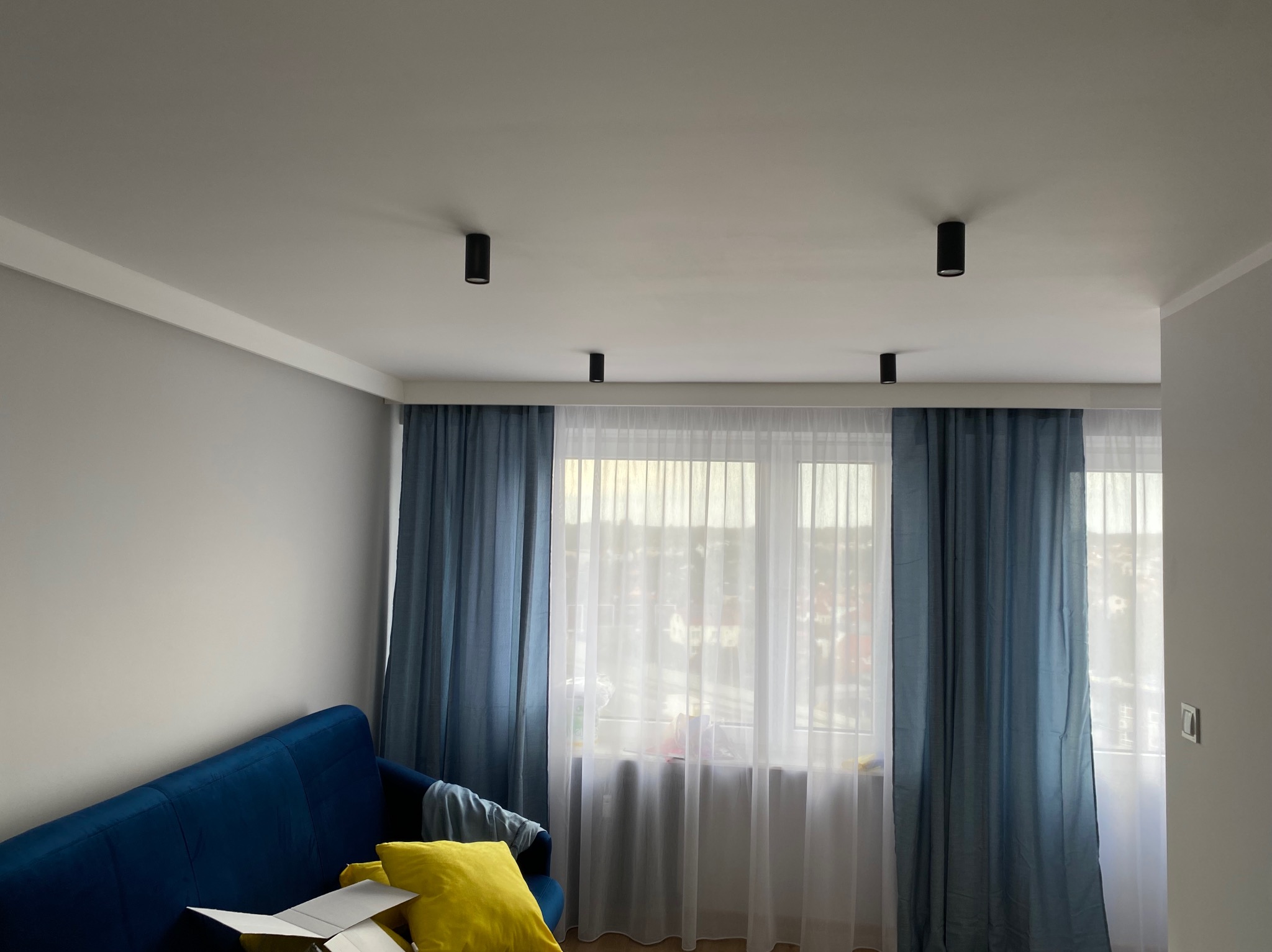 Wnętrze pokoju po odświeżeniu: szare ściany, biały sufit z czarnymi punktowymi lampami, błękitne zasłony i granatowa kanapa z żółtą poduszką.
