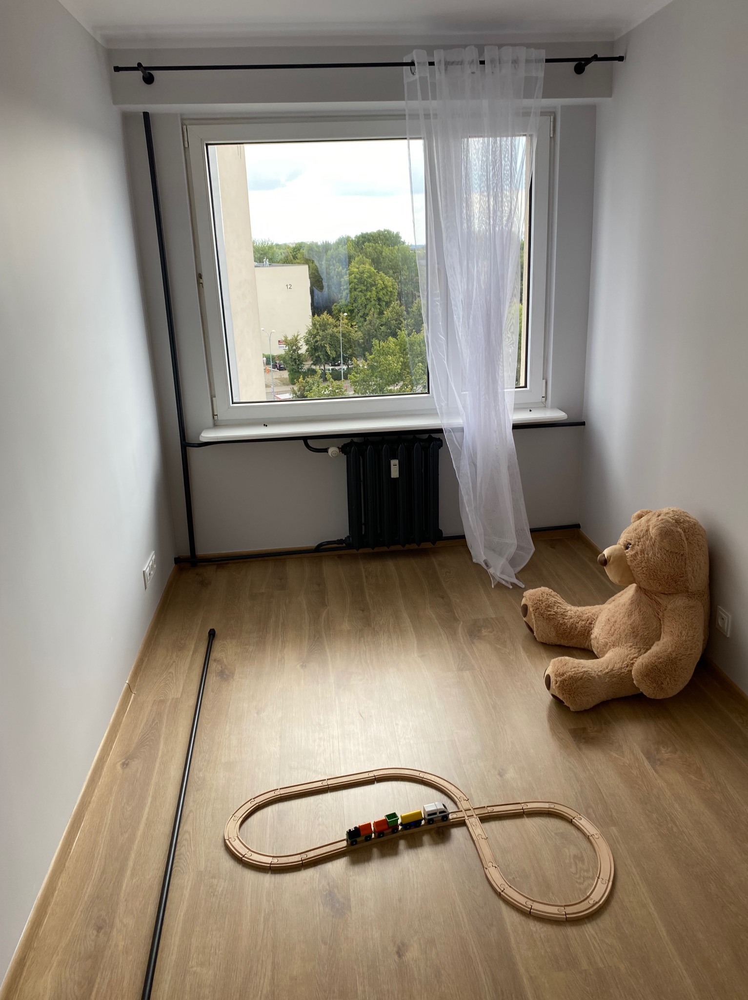 Pusty pokój po remoncie z drewnianą podłogą, białymi ścianami, czarnym grzejnikiem pod oknem z białą firanką, pluszowym misiem i drewnianą kolejką.