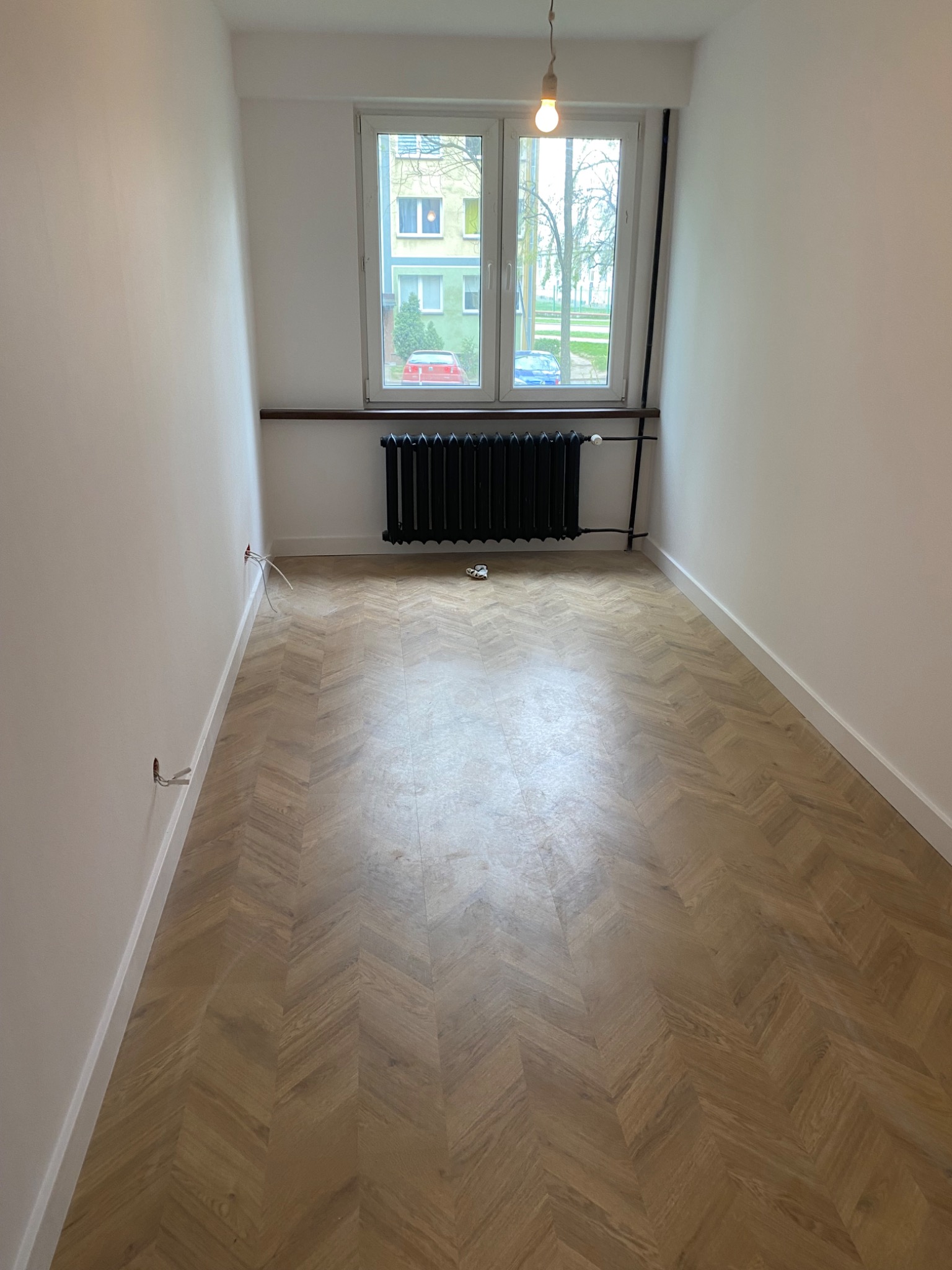 Puste, jasne pomieszczenie po remoncie z parkietem w jodełkę, białymi ścianami, grzejnikiem pod oknem i wiszącą żarówką, widoczne niezaizolowane kable w ścianie.