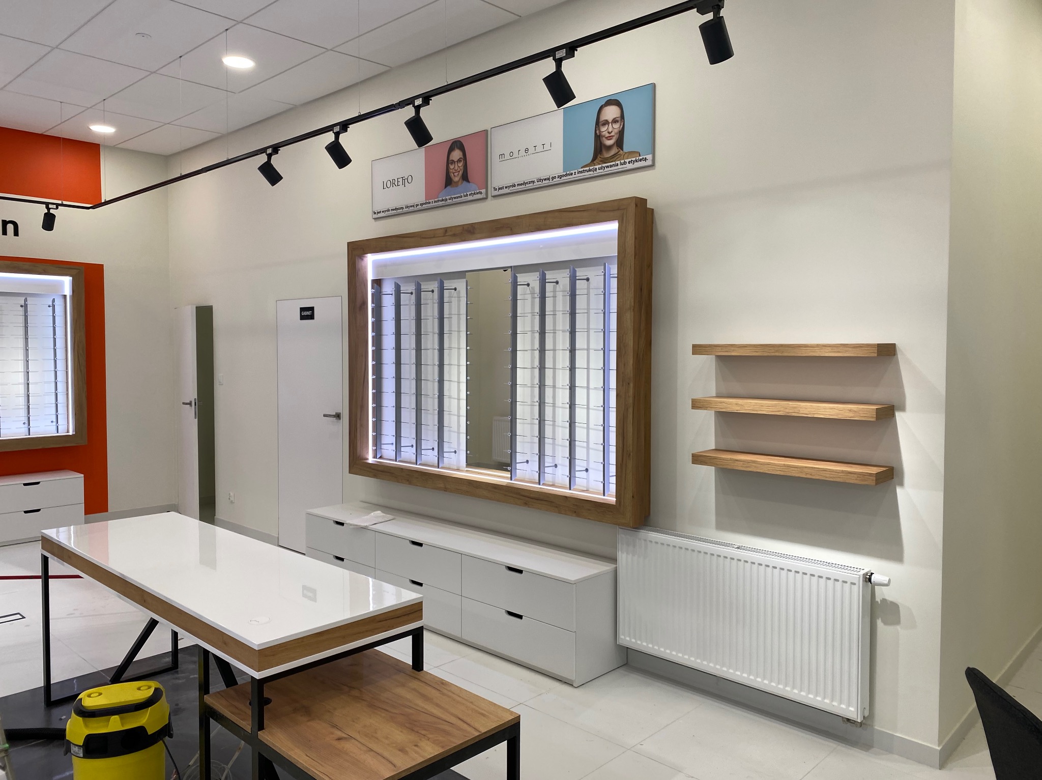 Wnętrze nowocześnie urządzonego salonu optycznego z białymi meblami, drewnianymi akcentami i oświetleniem szynowym, prezentujące ekspozycję okularów w podświetlanej ramie.