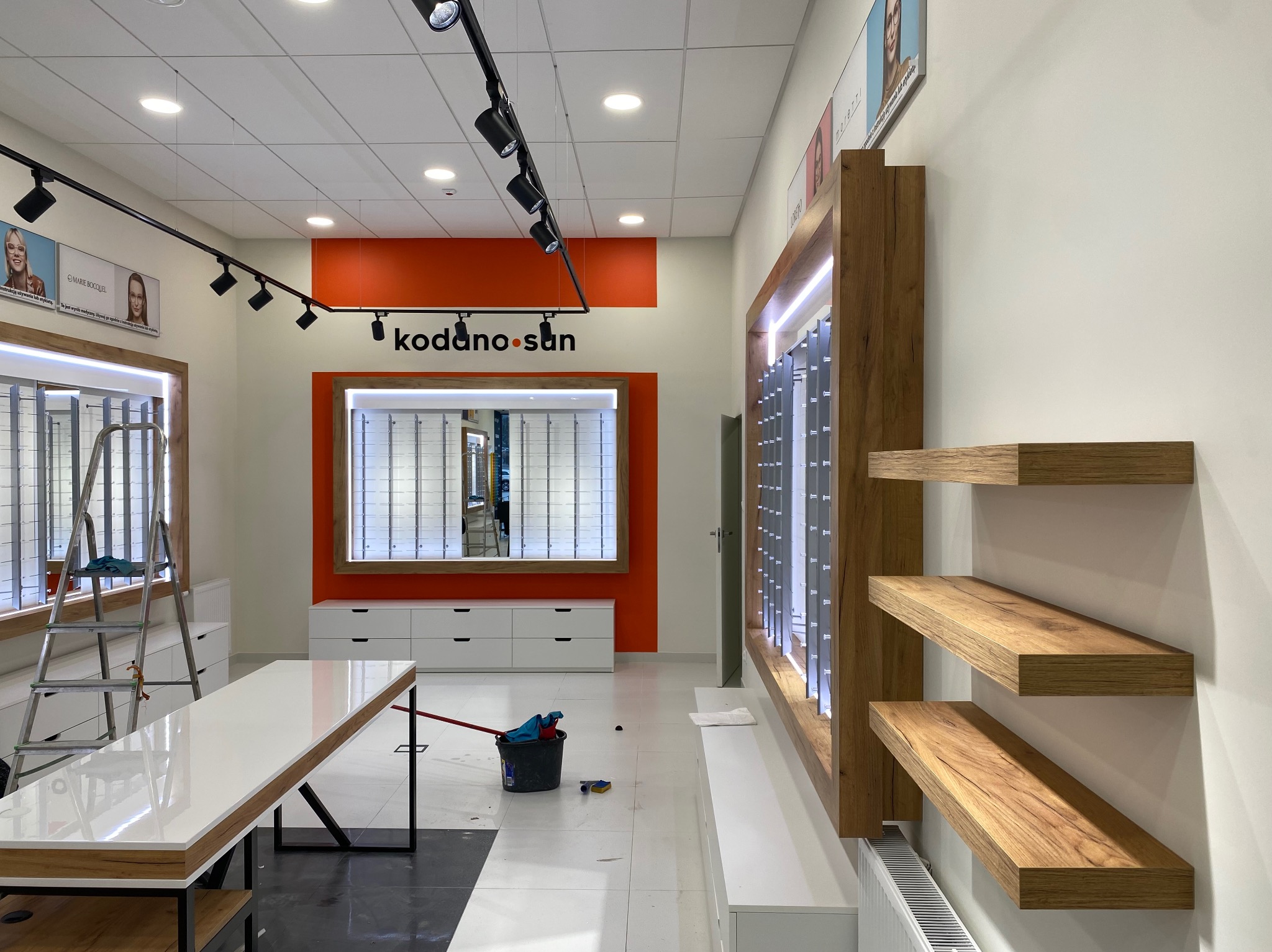 Wnętrze lokalu handlowego w trakcie sprzątania po remoncie: widoczne regały na okulary, drabina, wiadro z mopem i resztki farby na podłodze.