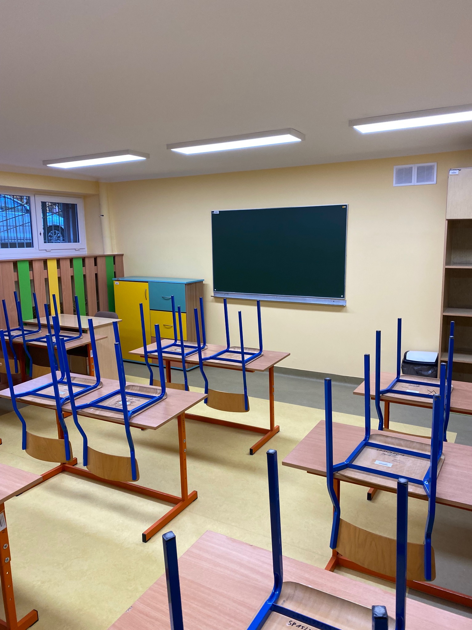 Pusta klasa szkolna z odwróconymi krzesłami na biurkach, ściany w jasnym odcieniu żółci, widoczna tablica i regał na książki, podłoga w kolorze piaskowym, oświetlenie sufitowe włączone.