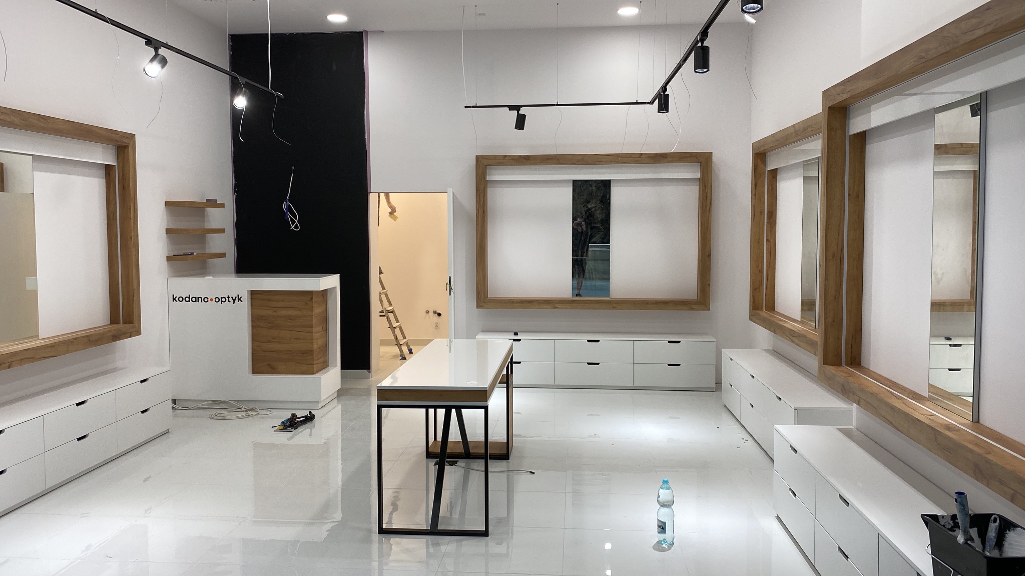 Wnętrze nowo wykończonego lokalu optycznego z białymi meblami z drewnianymi akcentami, widoczne narzędzia i butelka wody na błyszczącej podłodze, drabina w przejściu sugeruje trwające prace...