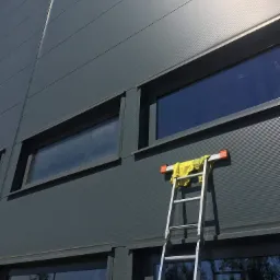 Aluminiowa drabina oparta o szarą ścianę budynku z widocznymi oknami; na szczycie drabiny leży żółta ścierka.