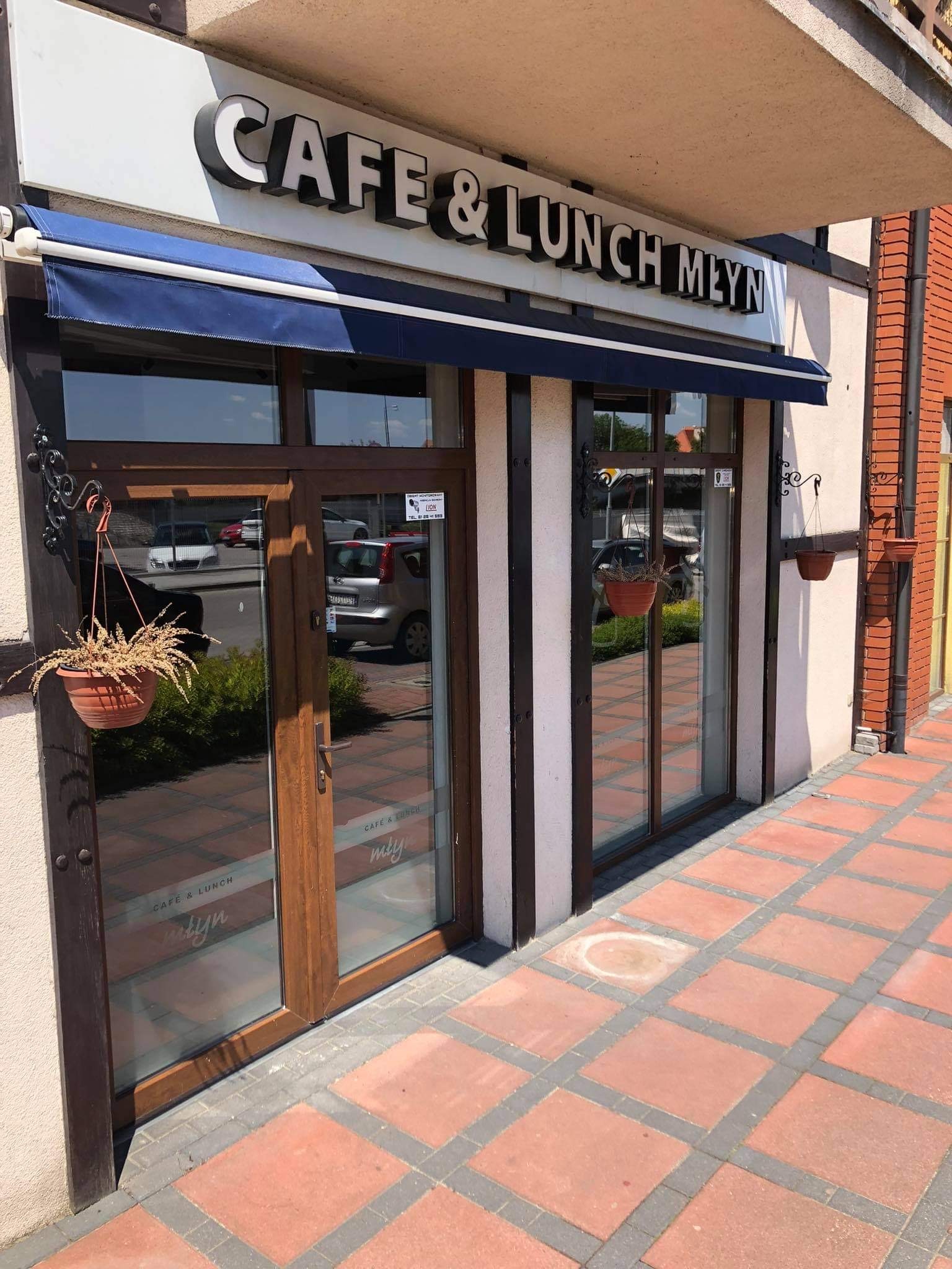 Wejście do kawiarni 'Cafe & Lunch Młyn' z ciemnymi ramami okiennymi i markizą, z wiszącymi doniczkami z kwiatami po bokach, odbicie ulicy i samochodów w szybach, oraz czerwona kostka brukowa przed...