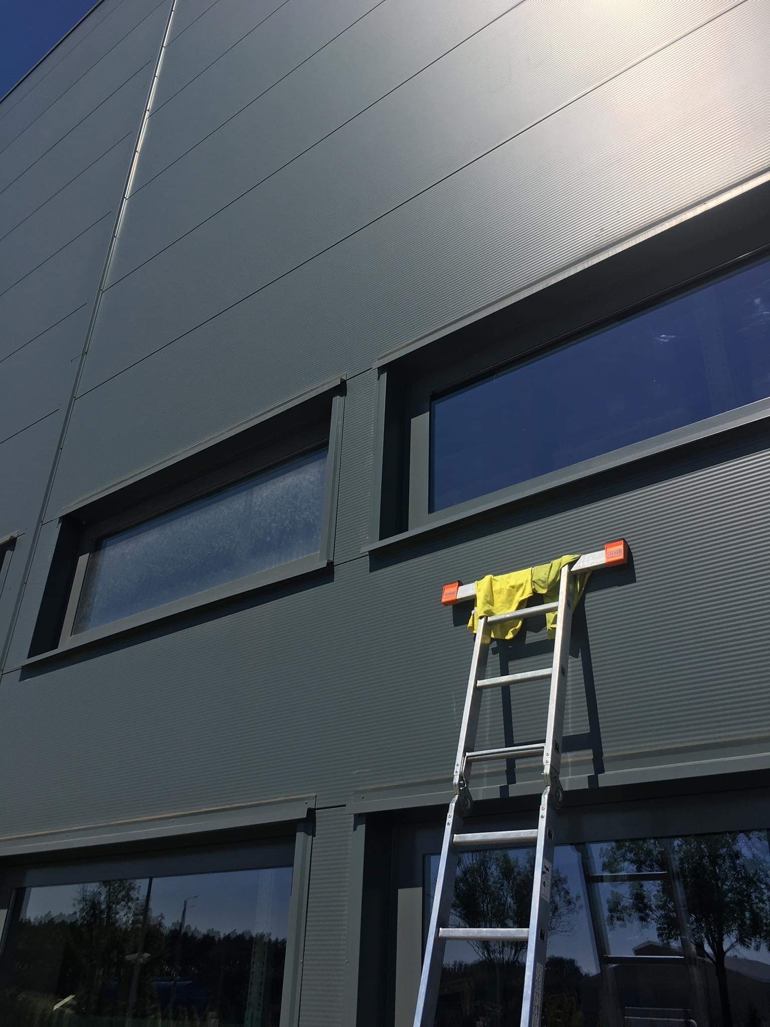 Aluminiowa drabina oparta o szarą ścianę budynku z widocznymi oknami; na szczycie drabiny leży żółta ścierka.