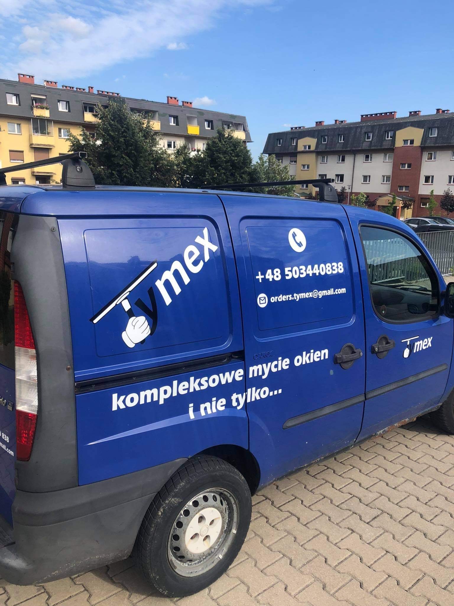 Niebieski samochód dostawczy z logo firmy oferującej kompleksowe usługi, w tym mycie okien, zaparkowany na kostce brukowej przed budynkiem mieszkalnym.