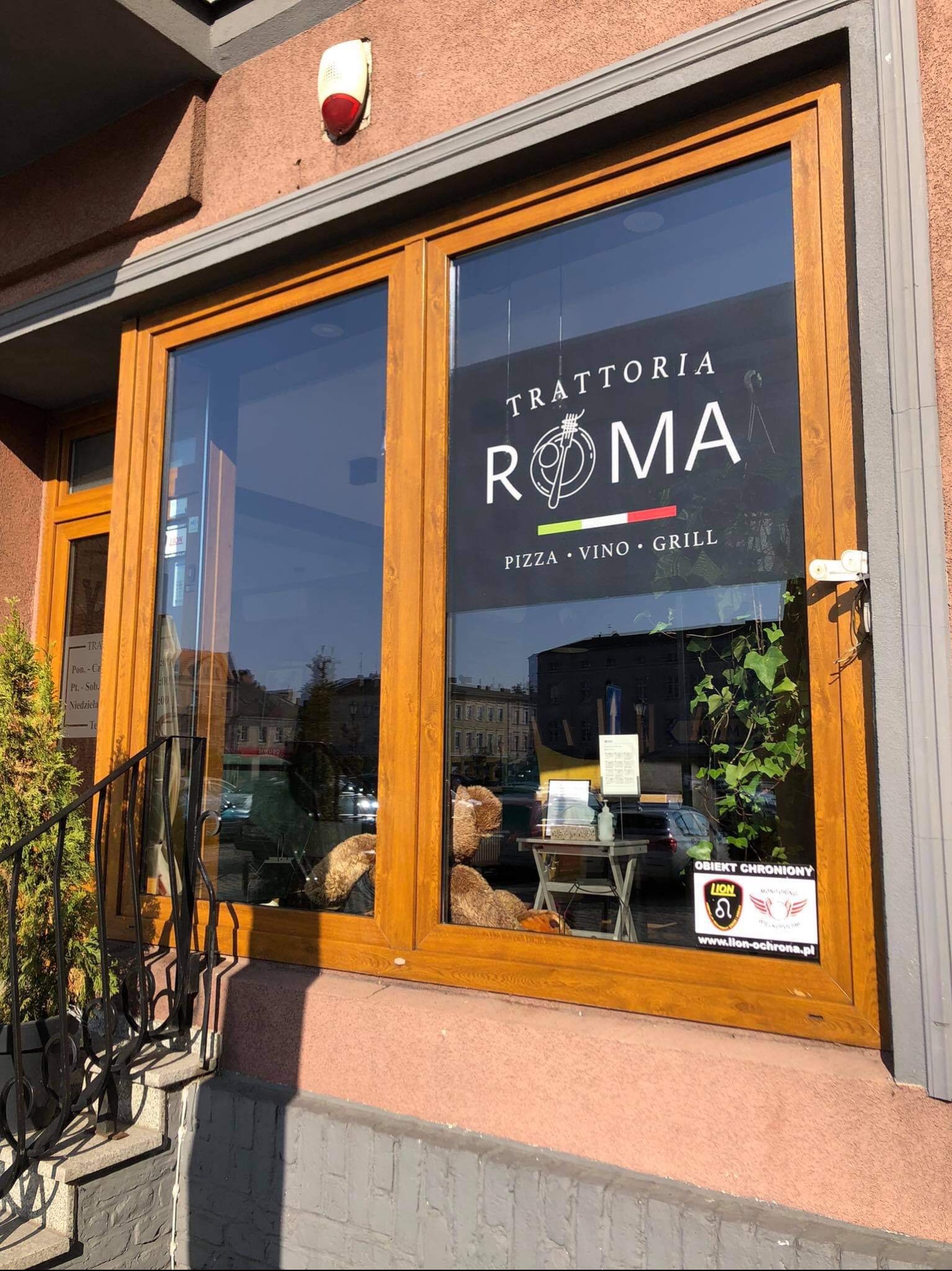 Fasada budynku z drewnianą witryną restauracji Trattoria Roma, widoczny stolik z dekoracjami wewnątrz, naklejka informująca o ochronie obiektu oraz mała choinka przy schodach.