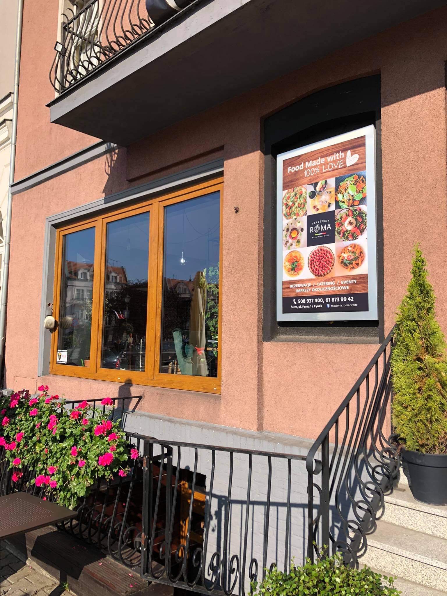 Fasada budynku z oknem i plakatem reklamowym restauracji Trattoria Roma, kwiaty w donicach i ozdobne barierki