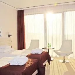 Jasny pok&oacute;j hotelowy z dwoma ł&oacute;żkami pojedynczymi, białymi szlafrokami, nowoczesnymi krzesłami i obrazem z motywem morskim na ścianie, oświetlony promieniami słonecznymi wpadającymi przez okno.