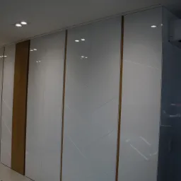 Biała szafa z przesuwnymi drzwiami, zdobiona delikatnym geometrycznym wzorem, z elementami drewnianymi, oświetlona punktowymi lampami sufitowymi. Na podłodze widoczny zielony pojemnik z akcesoriami.