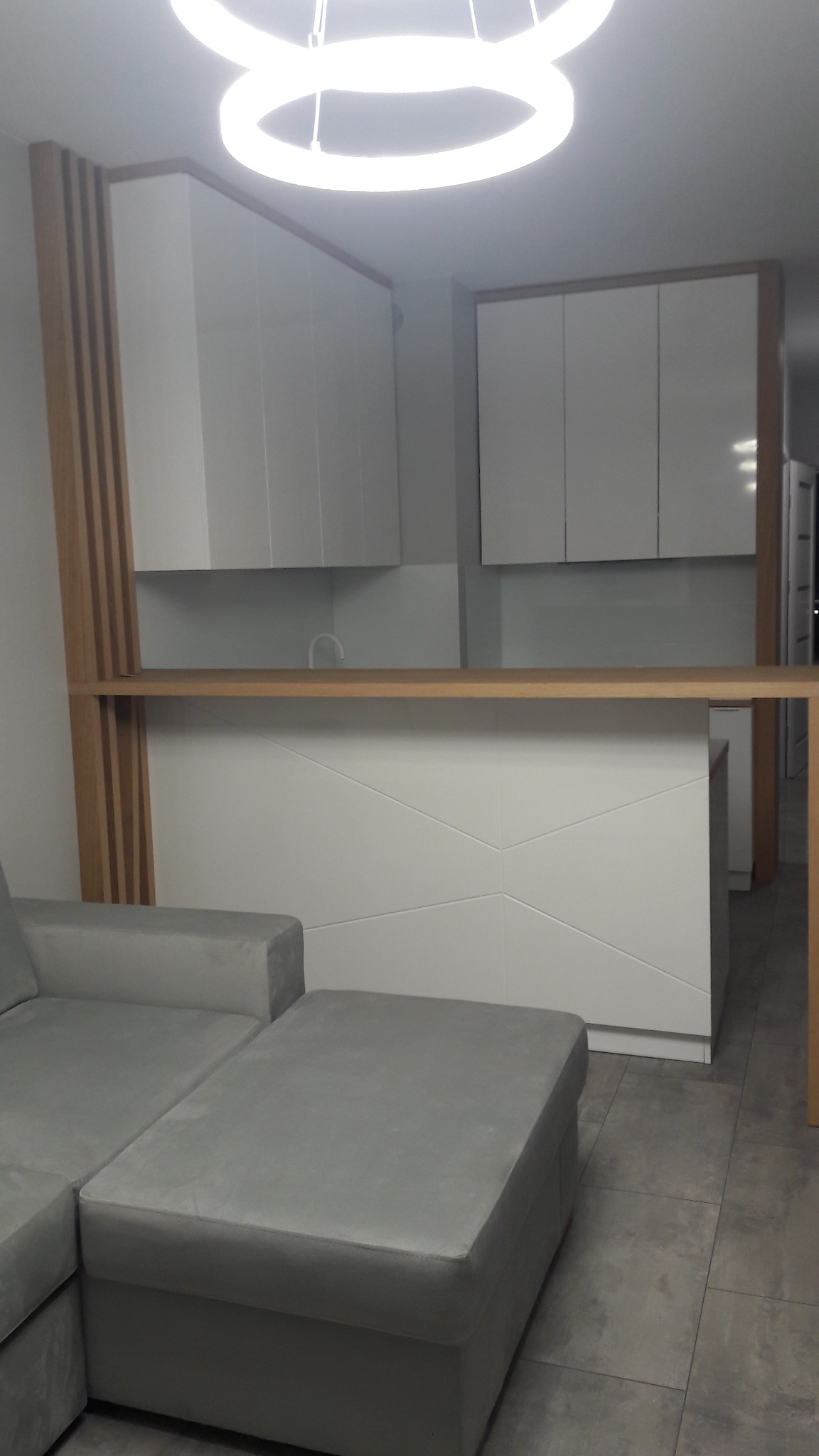 Wnętrze z białymi szafkami kuchennymi podwieszanymi i wyspą z geometrycznym wzorem, oddzielone drewnianą konstrukcją od szarej sofy modułowej, oświetlone nowoczesną lampą wiszącą w kształcie...