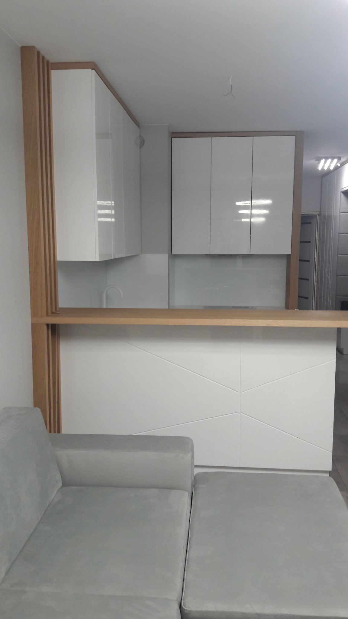 Nowoczesna kuchnia z białymi szafkami, drewnianym blatem i elementami dekoracyjnymi z drewna, widoczny fragment szarej sofy.