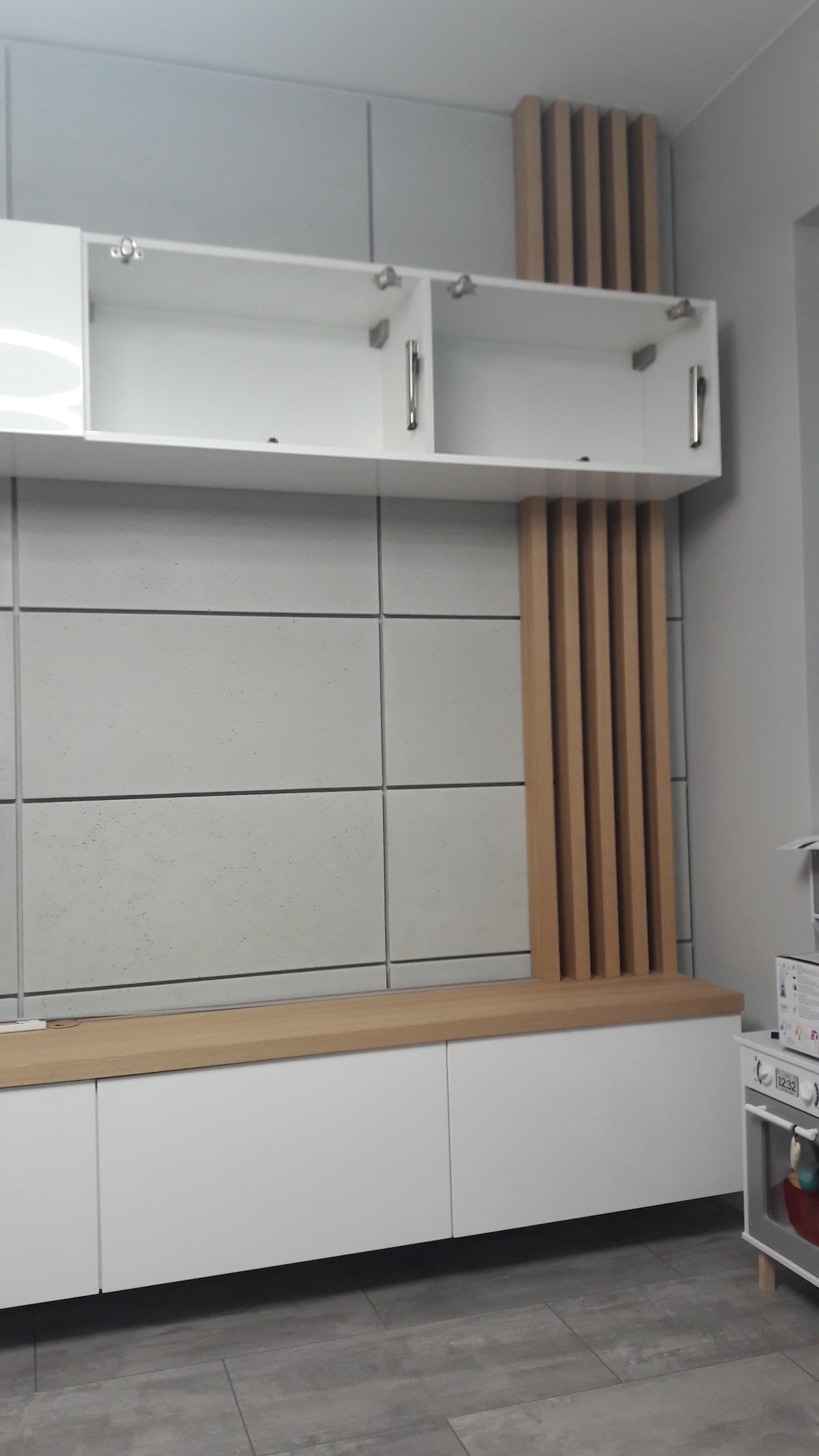 Białe szafki wiszące i stojące z drewnianymi akcentami w postaci pionowych listew, na tle ściany z szarych płyt, widoczna dziecięca kuchenka.