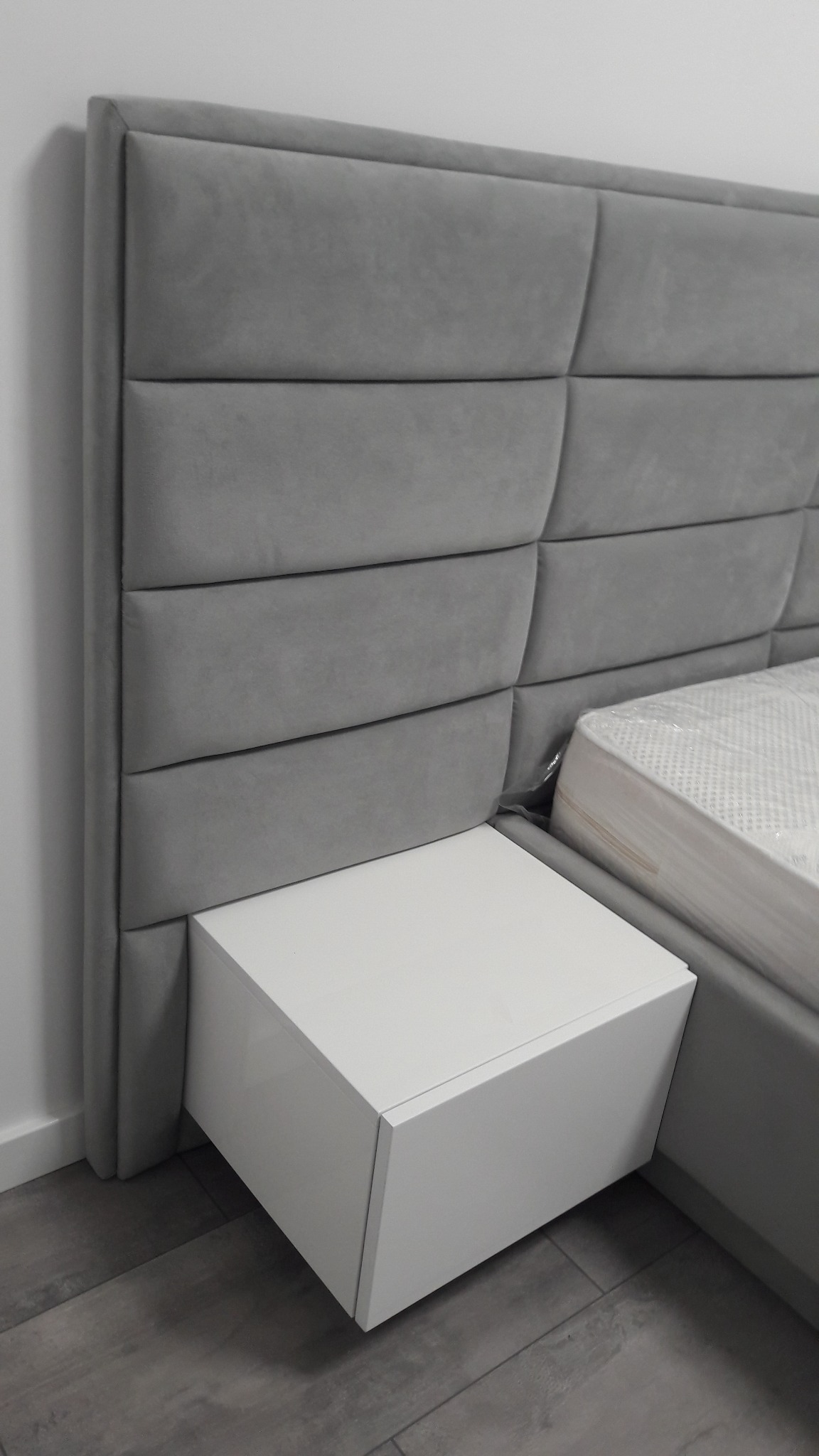 Szare, tapicerowane wezgłowie łóżka z geometrycznym wzorem, biała szafka nocna z wysuwaną szufladą, materac w folii ochronnej.