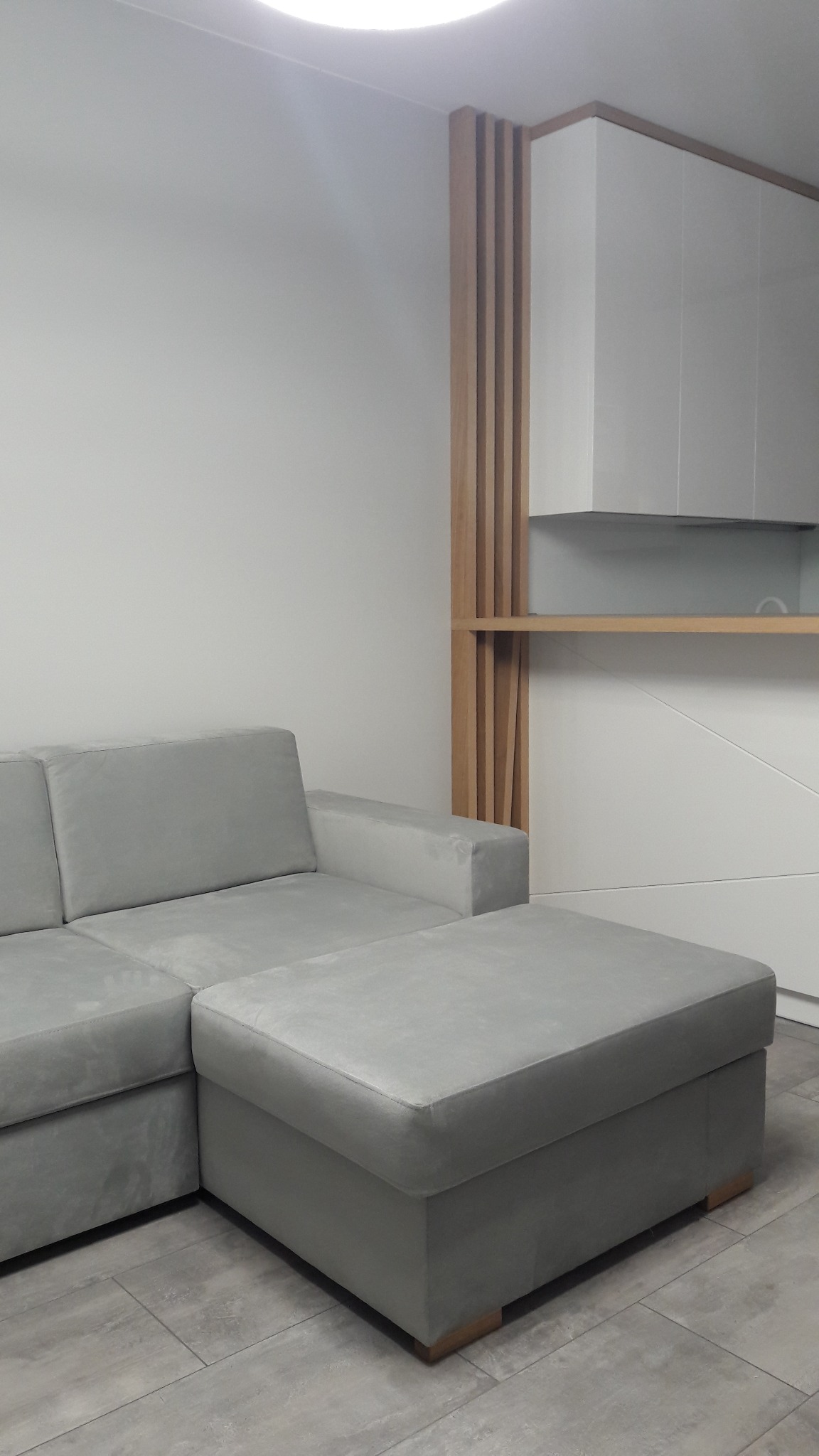 Szary narożnik z pufą na tle białej zabudowy kuchennej z drewnianymi akcentami, szare płytki podłogowe.