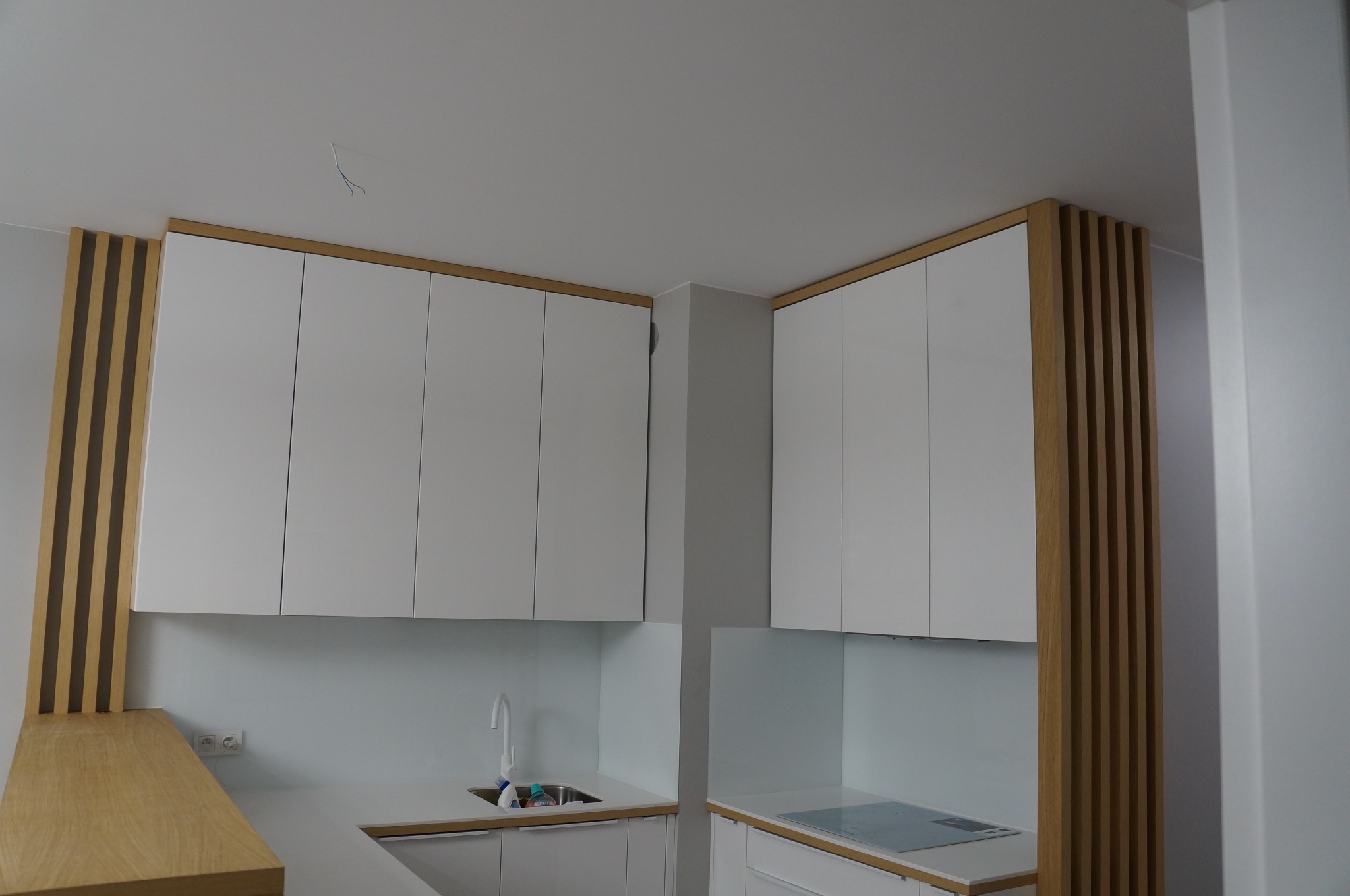 Nowoczesna kuchnia z białymi szafkami wiszącymi, drewnianymi akcentami w postaci pionowych listew i blatem z jasnego drewna, minimalistyczny design, widoczny fragment zlewu i płyty indukcyjnej.