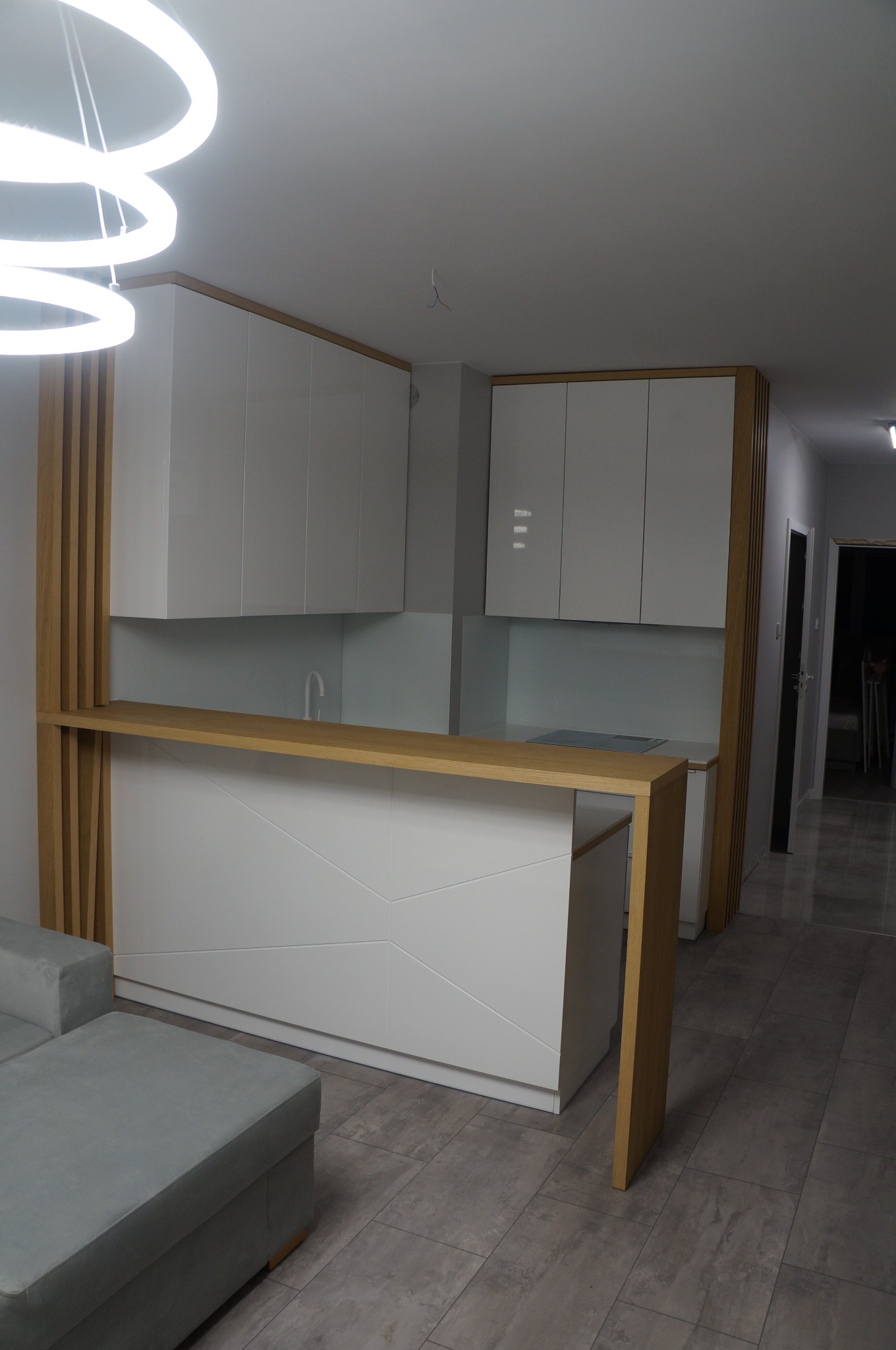 Nowoczesna kuchnia z białymi szafkami i drewnianym blatem w Kielcach, widoczna z perspektywy salonu, z sofą w kolorze gołębiej szarości i geometrycznym wzorem na frontach.