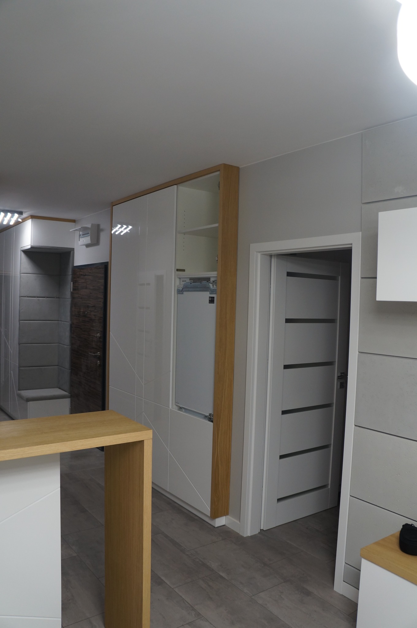 Wnętrze mieszkania z zabudową szaf z białymi, lakierowanymi frontami i drewnianymi obramowaniami, widoczna również zabudowa przedpokoju z tapicerowanym siedziskiem oraz drzwi wewnętrzne z poziomymi...