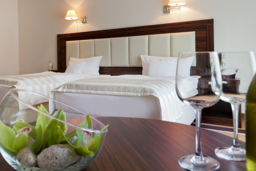 Pokój hotelowy z dwoma łóżkami pojedynczymi, ciemnobrązowym wezgłowiem z beżowymi panelami, stolik z dekoracją z zielonych orchidei i kamieni w szklanej misie oraz dwa kieliszki do wina...