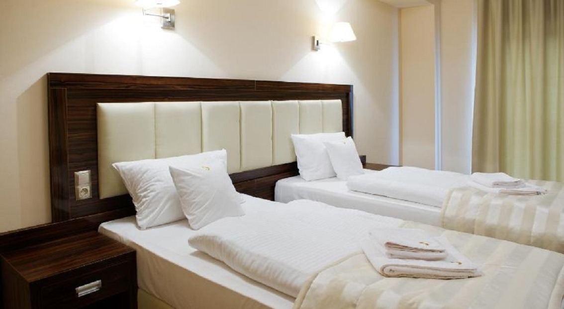 Pokój hotelowy z dwoma pojedynczymi łóżkami, ciemnobrązowe wezgłowie z panelami w kolorze kremowym, biała pościel z ręcznikami, nocny stolik z gniazdkiem elektrycznym.