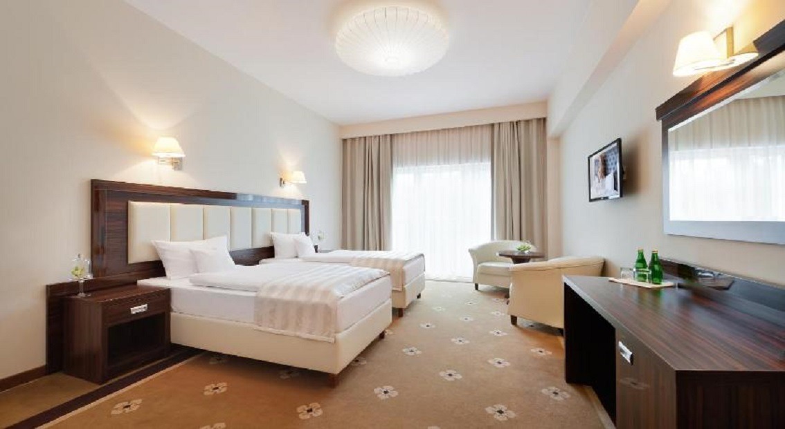 Jasny pokój hotelowy z dwoma pojedynczymi łóżkami, ciemnobrązowymi szafkami nocnymi i biurkiem, kremowym fotelem oraz telewizorem na ścianie.