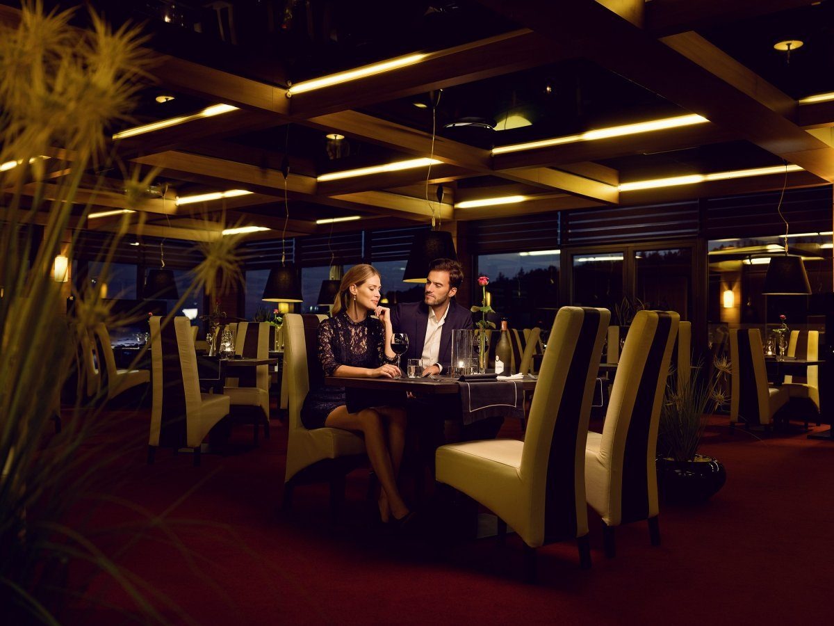 Eleganckie wnętrze restauracji z parą siedzącą przy stoliku, oświetlenie punktowe podkreśla drewniane belki sufitowe, krzesła tapicerowane w beżowo-brązowe pasy, czerwona wykładzina.