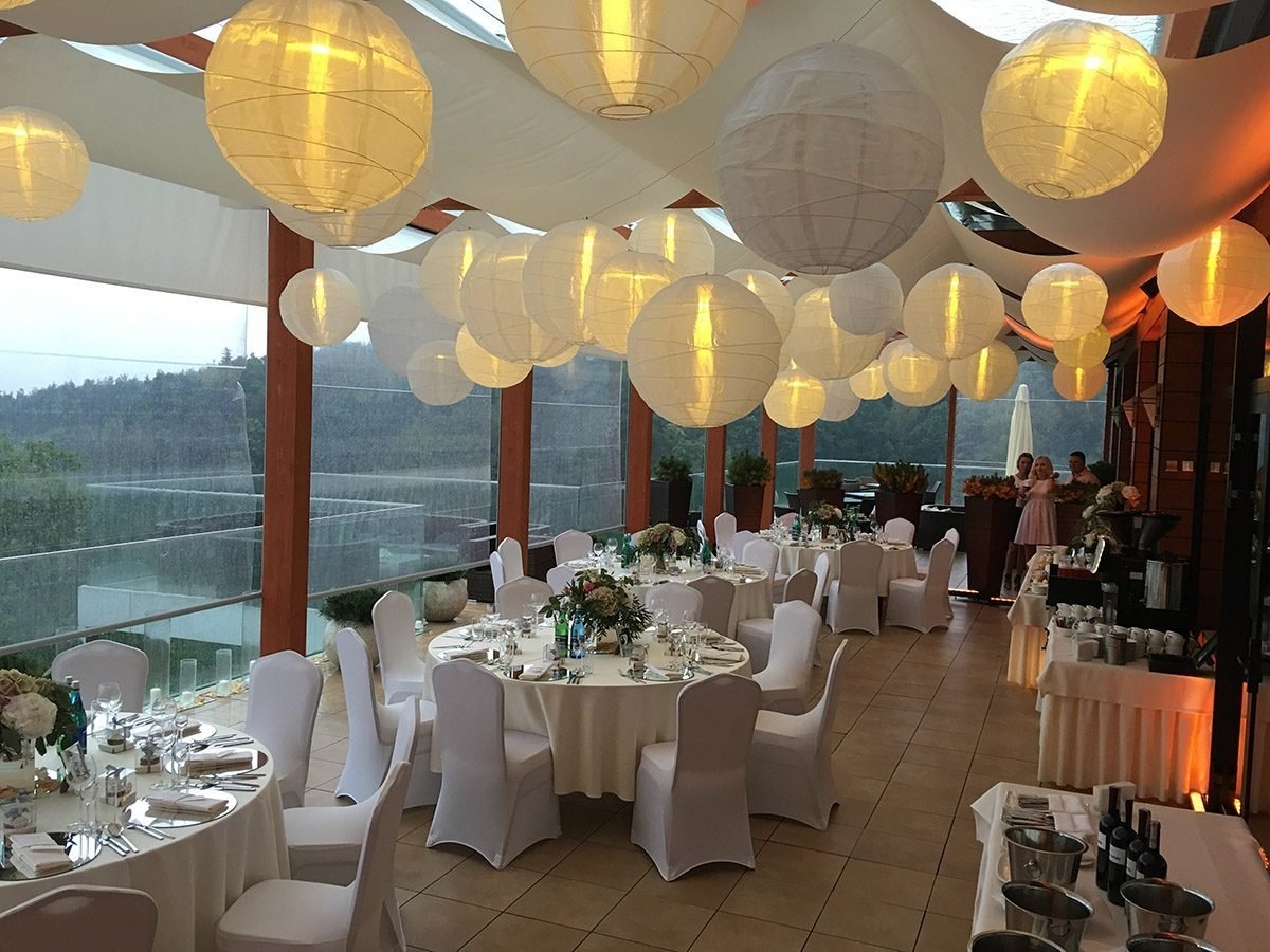 Uroczysta sala bankietowa z okrągłymi stołami nakrytymi białymi obrusami i krzesłami w pokrowcach, udekorowana kwiatami i lampionami, widok na zielony krajobraz za oknem.