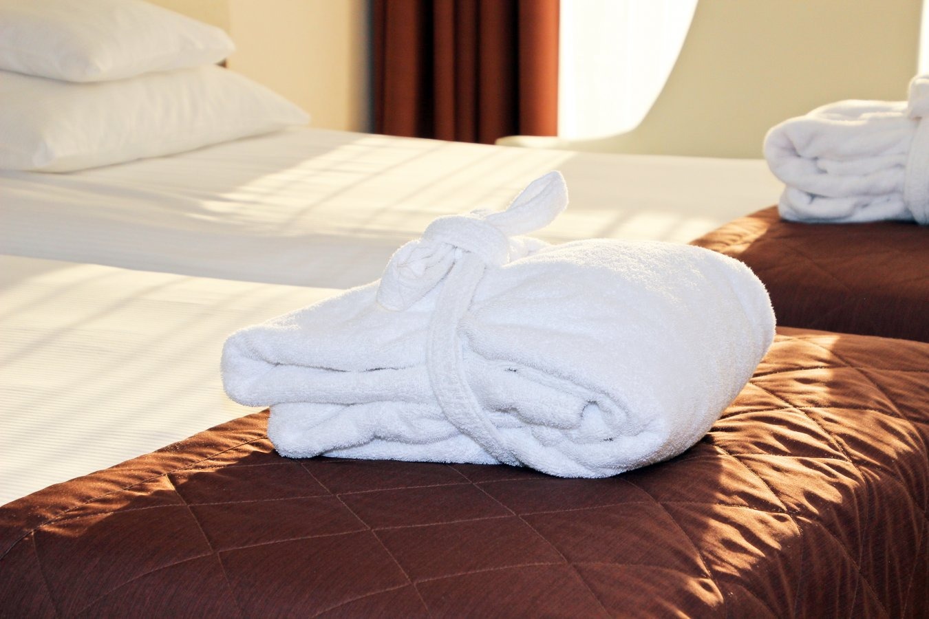 Dwa łóżka hotelowe z białymi ręcznikami i brązowymi narzutami z pikowaniem w romby, widok z bliska na złożony ręcznik z pętelką.